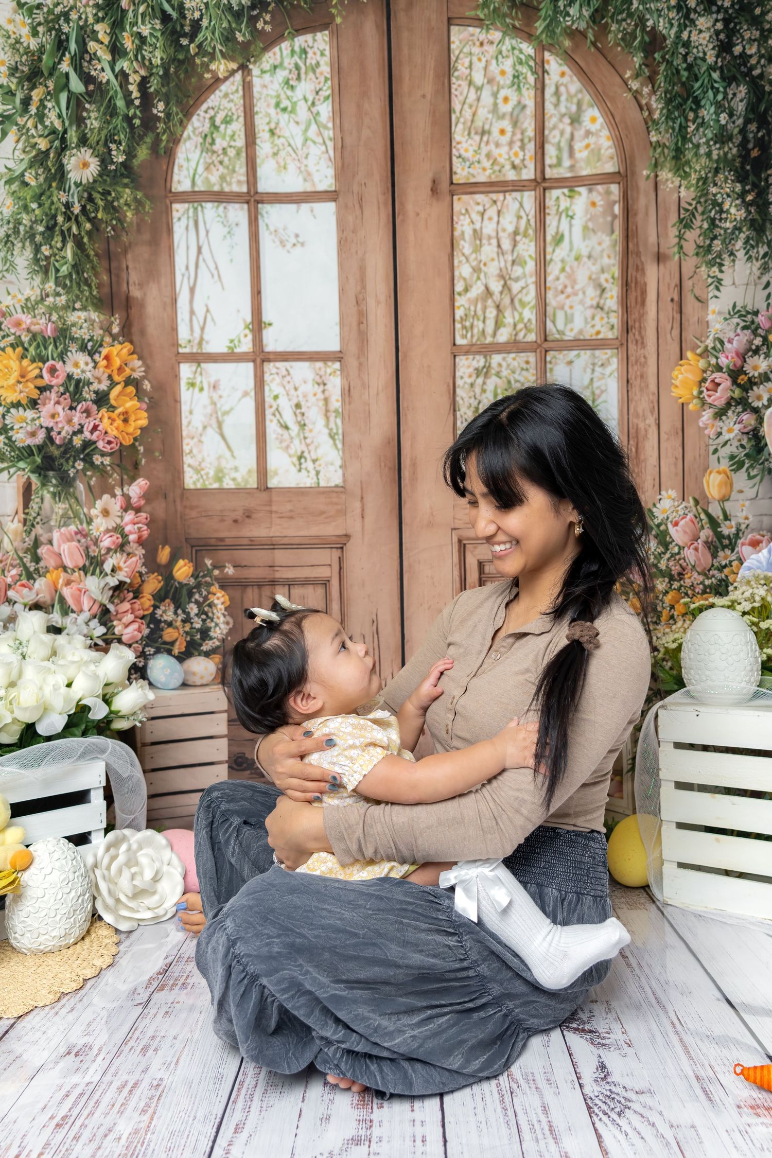 A smiling mother sits on a white wooden floor holding her baby girl, surrounded by colorful flowers and a rustic wooden door backdrop.