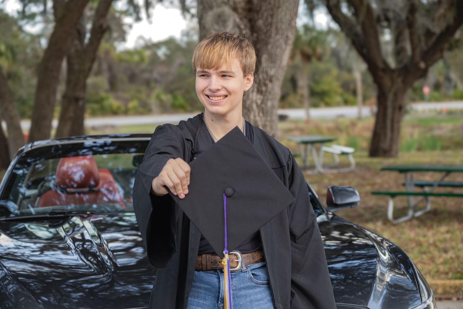 Smiling graduate in black gown holds cap outdoors near a black car, celebrating graduation day.