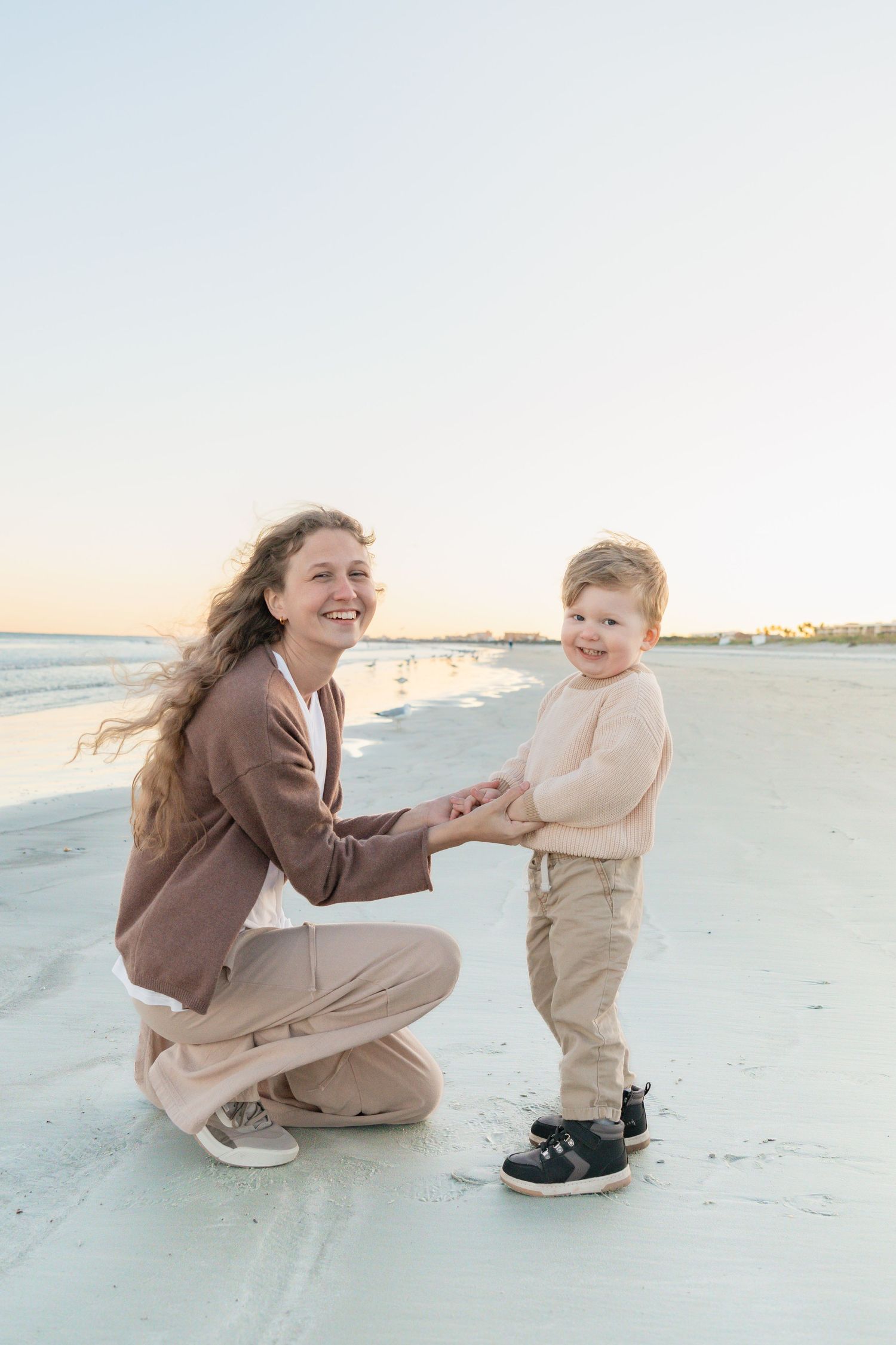 A smiling woman crouches on a sandy beach holding hands with a happy toddler boy at sunset.