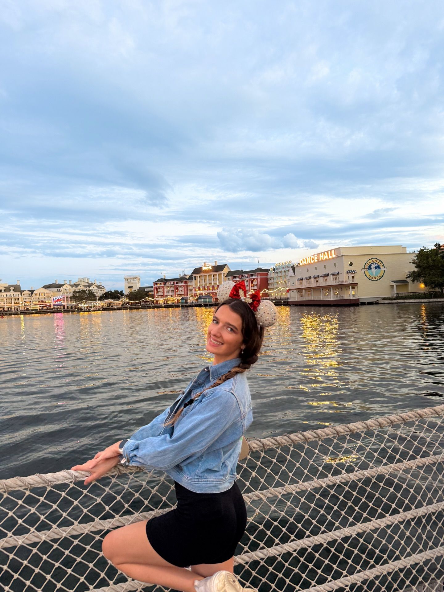 Smiling woman wearing Mickey Mouse ears sitting on fence railing with Disney resort waterfront behind her.