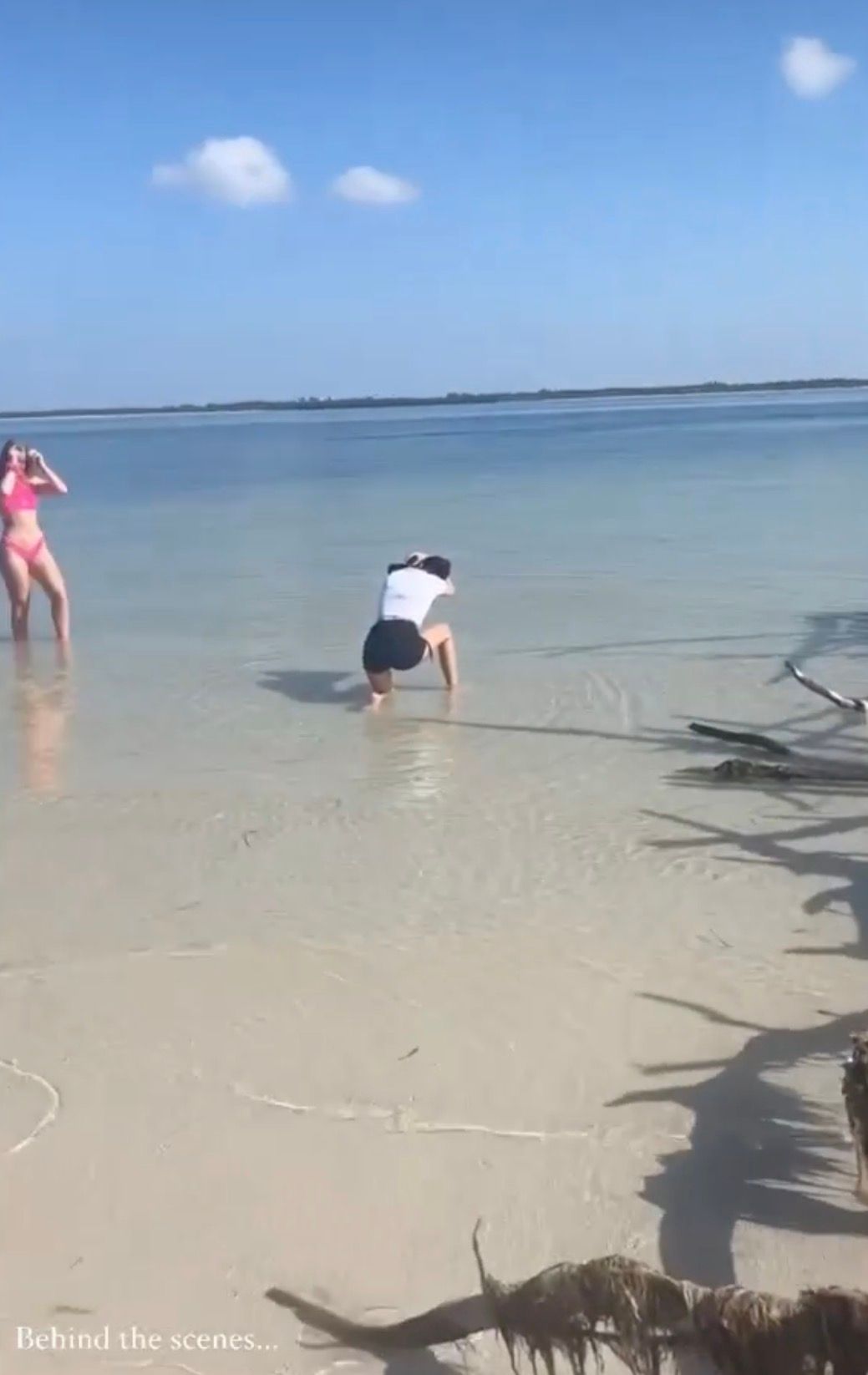 Behind-the-scenes beach photoshoot with photographer crouching in shallow water while woman in pink bikini poses nearby.
