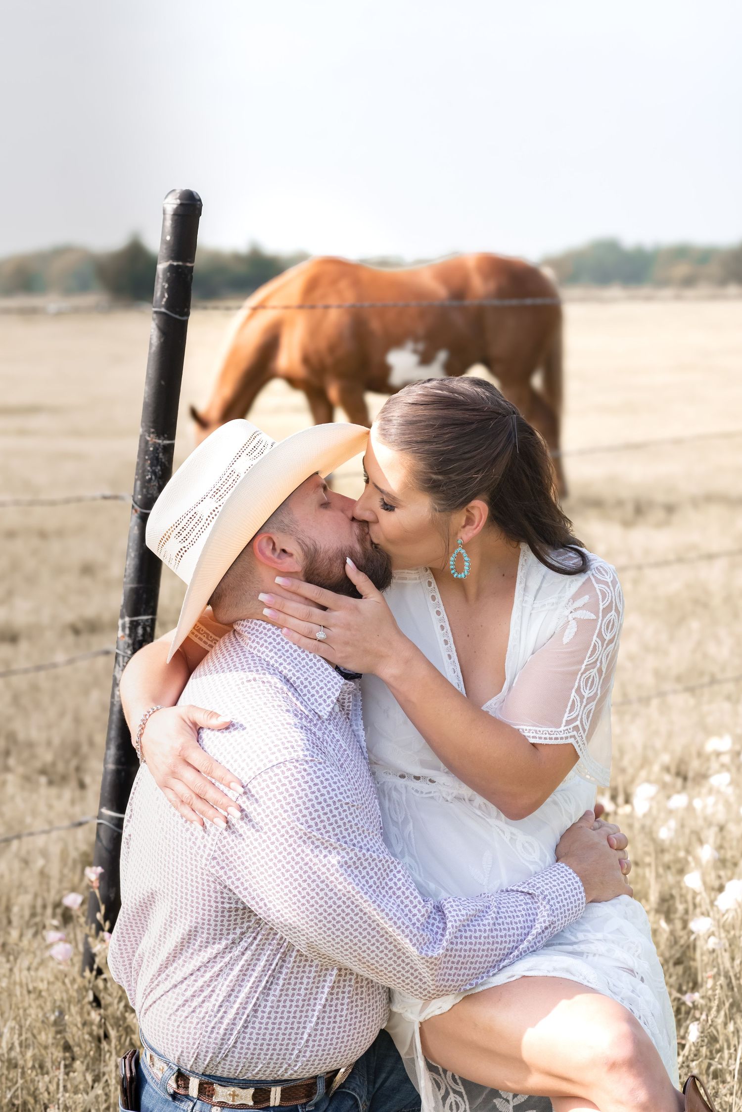 Couple kissing by fence post in golden field with horse grazing in background.