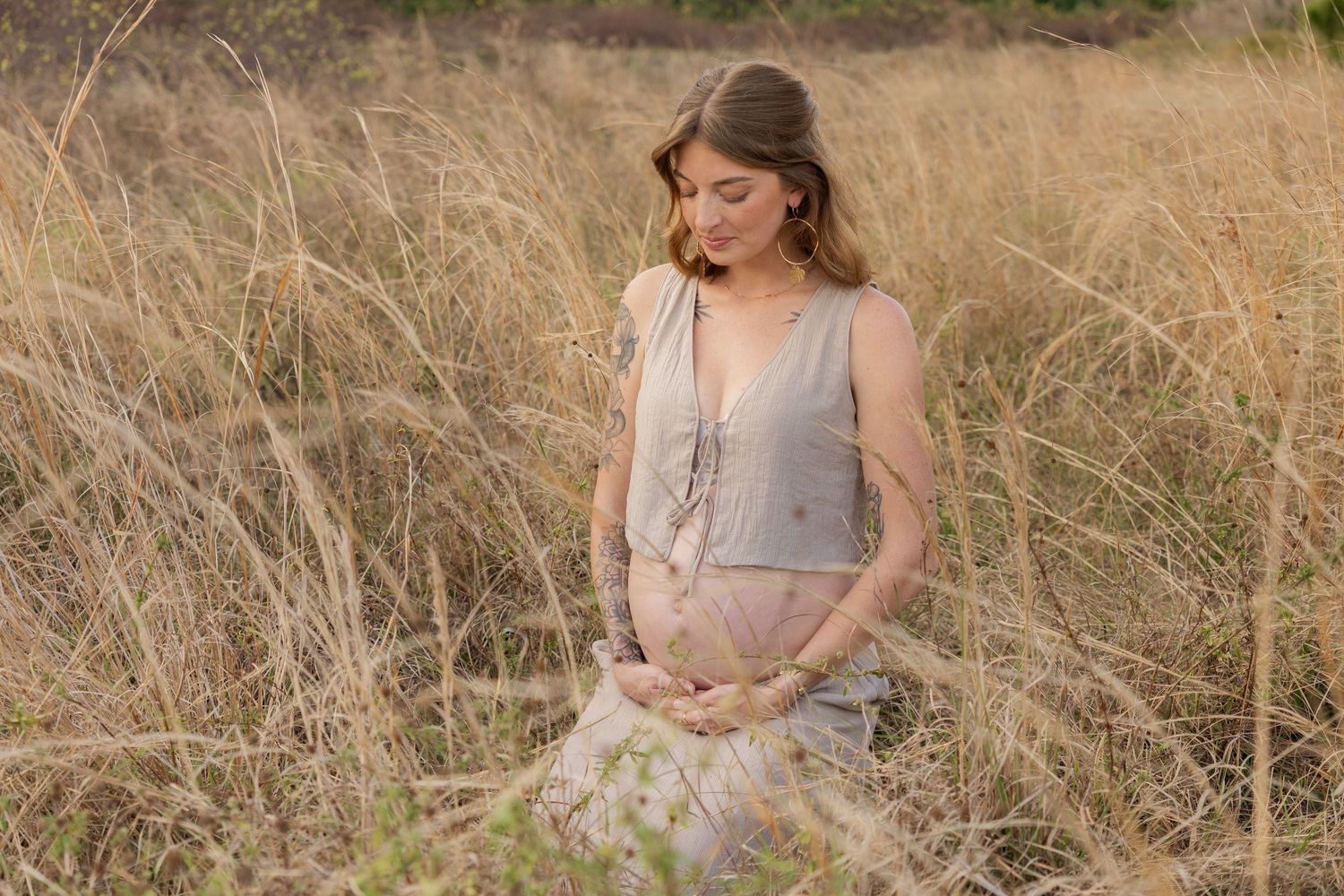Pregnant woman in gray dress sitting in tall golden grass field during maternity photo session.