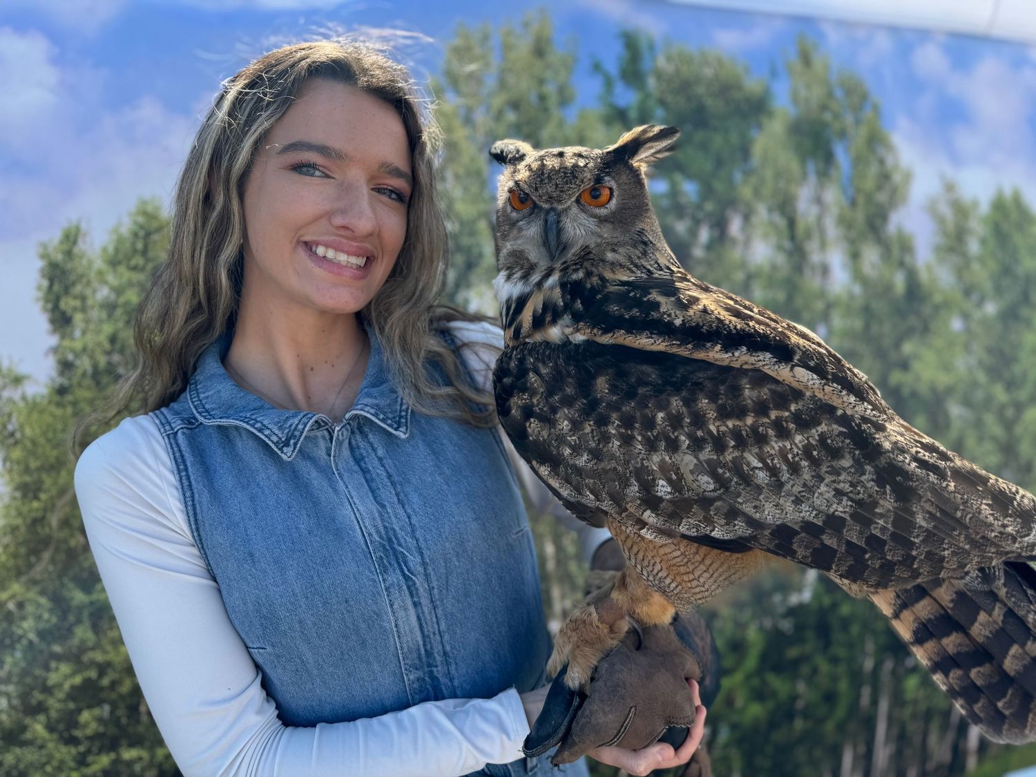 A smiling woman holds a large great horned owl on her gloved hand outdoors.