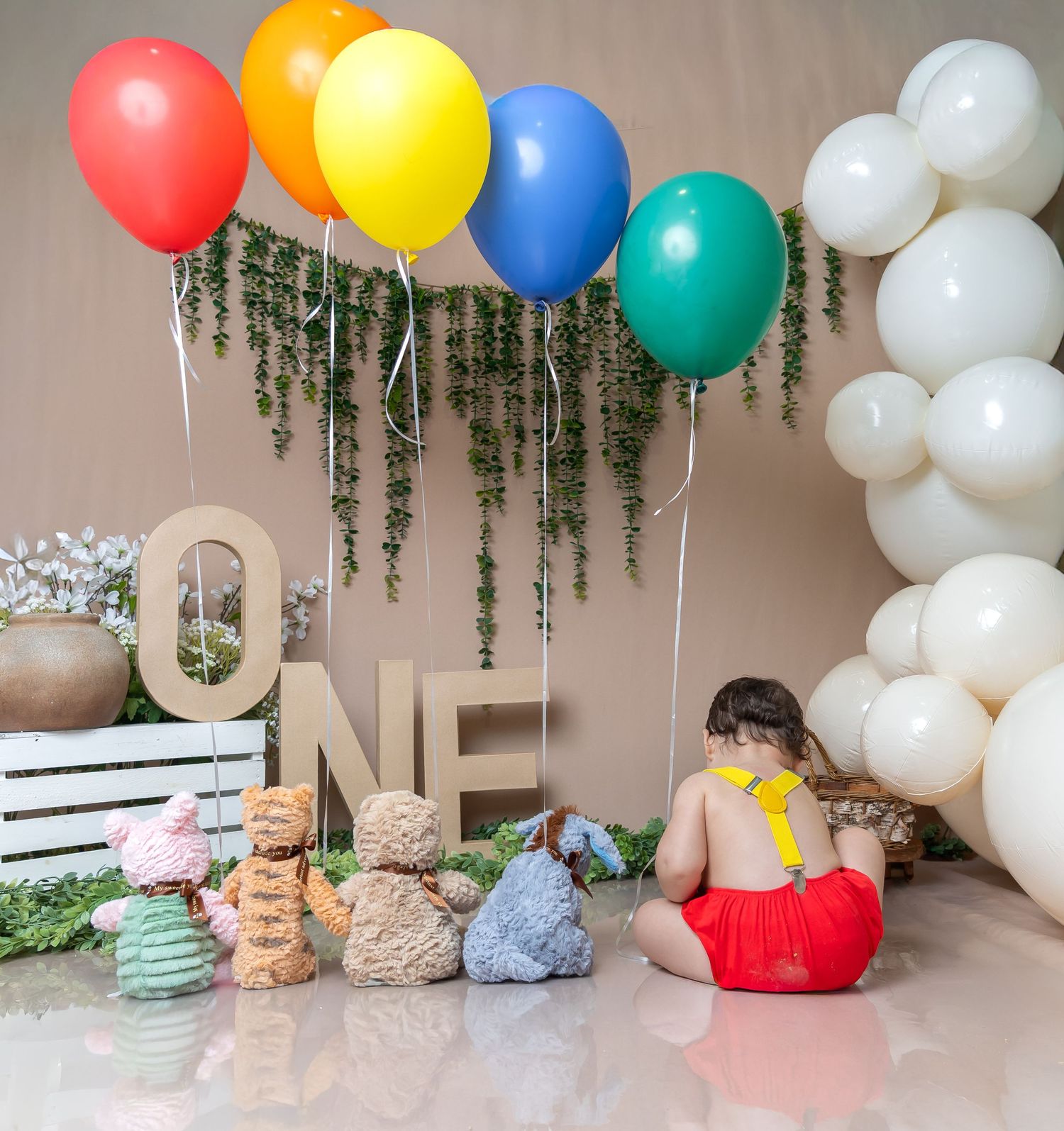 Baby's first birthday setup with colorful balloons, ONE letters, teddy bears, and a child in red overalls.