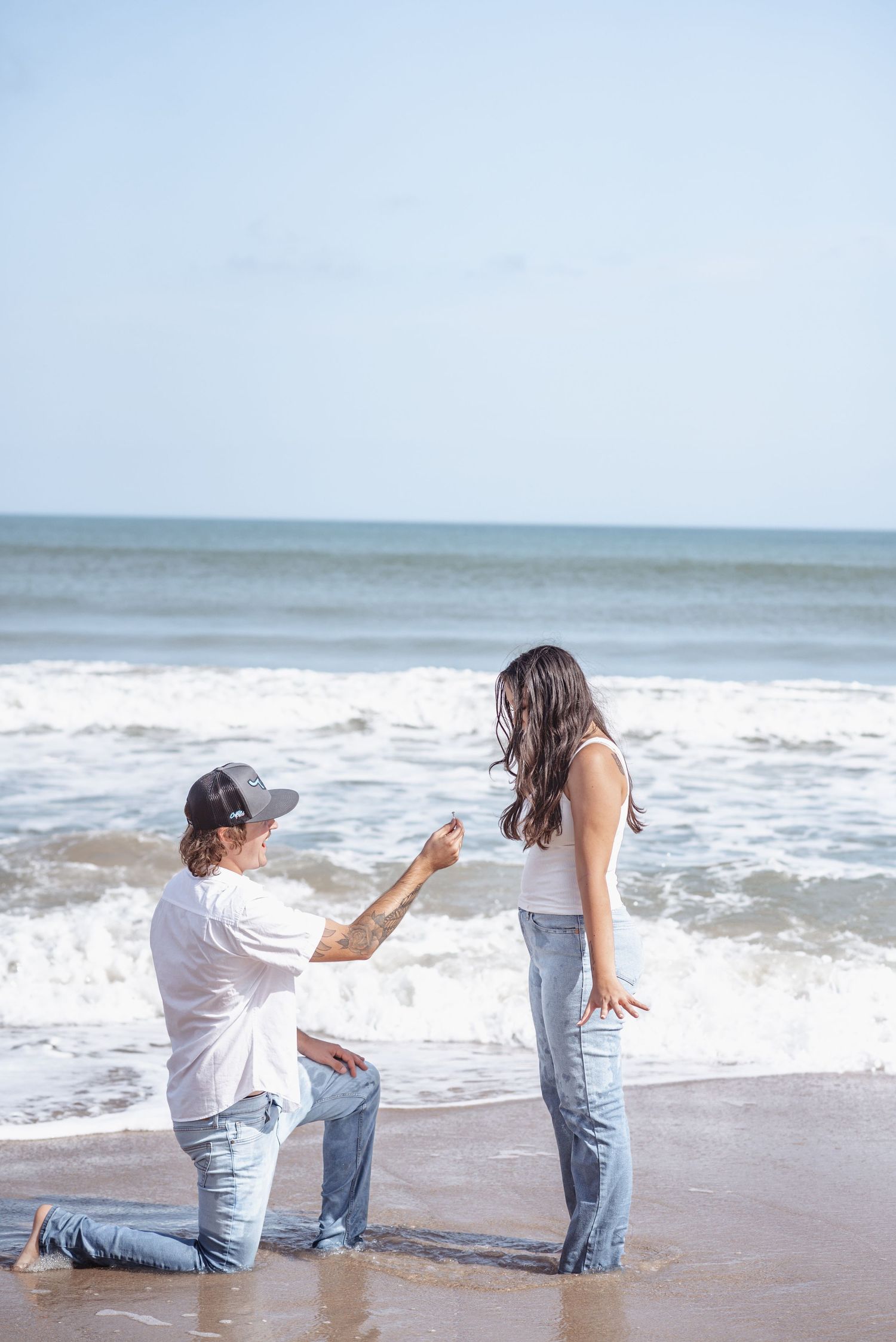 Man proposes on one knee to woman on a sunny beach with ocean waves in the background.