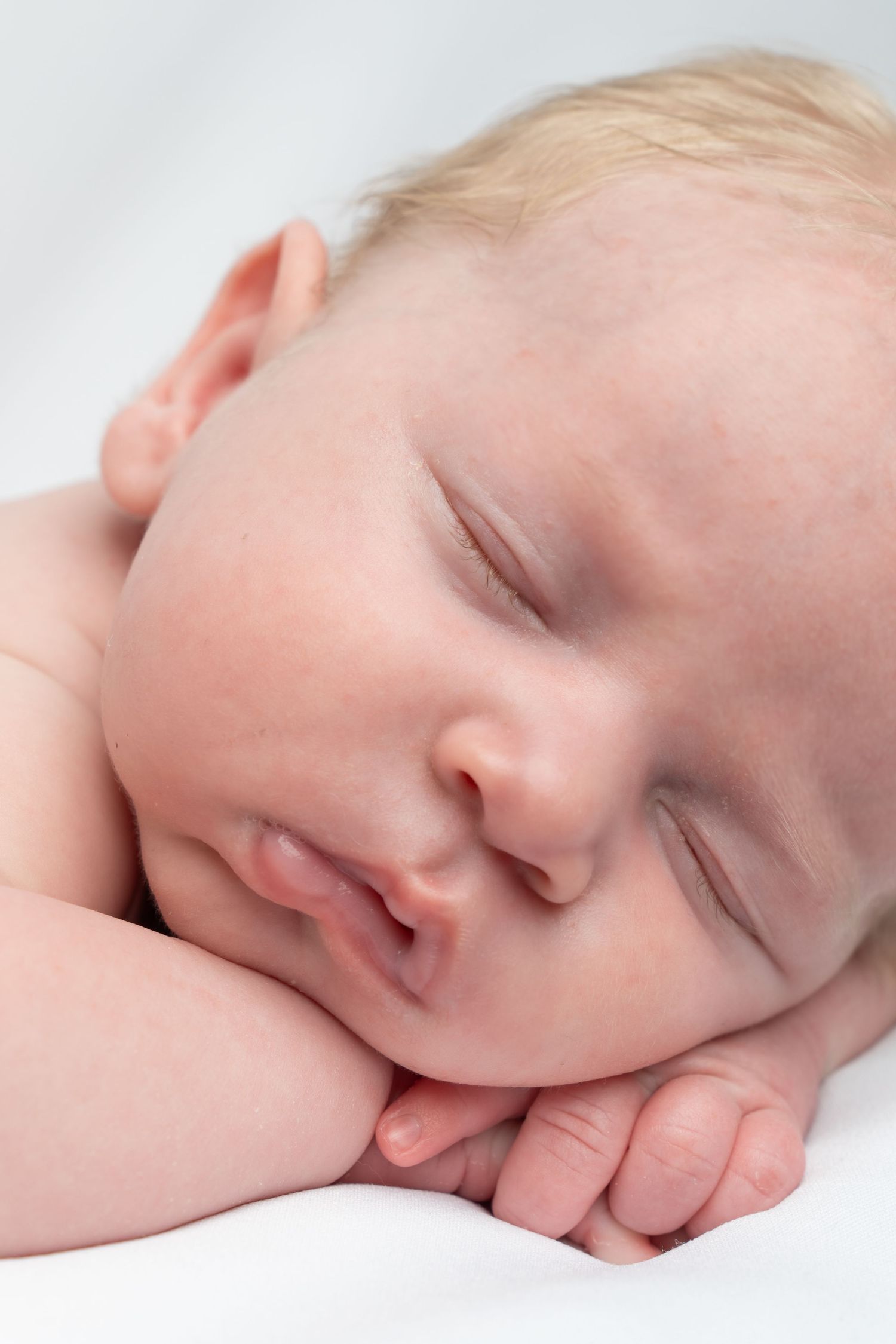 Close-up of a peaceful sleeping newborn baby with blonde hair resting on folded hands against white background.