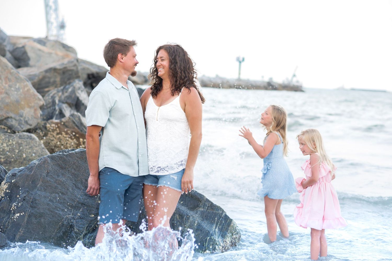 Family of four enjoying a beach day, parents laughing while two young girls play in the ocean waves near rocky shore.