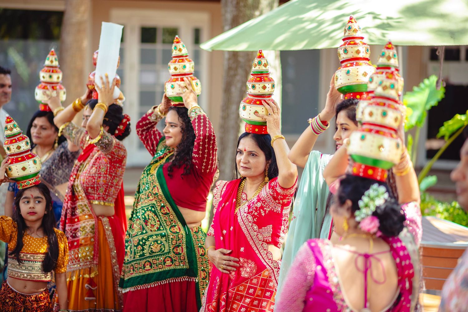 Women in vibrant Indian attire balance decorated pots on their heads during a traditional Hindu wedding ceremony outdoors.