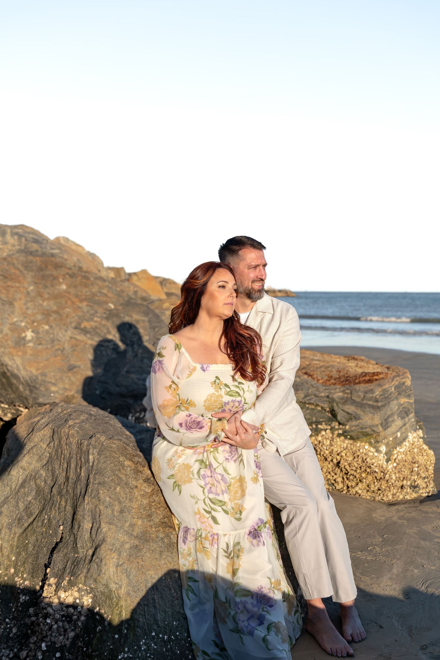 Couple embracing on a rocky beach at sunset, woman in floral dress leaning against man in linen suit.