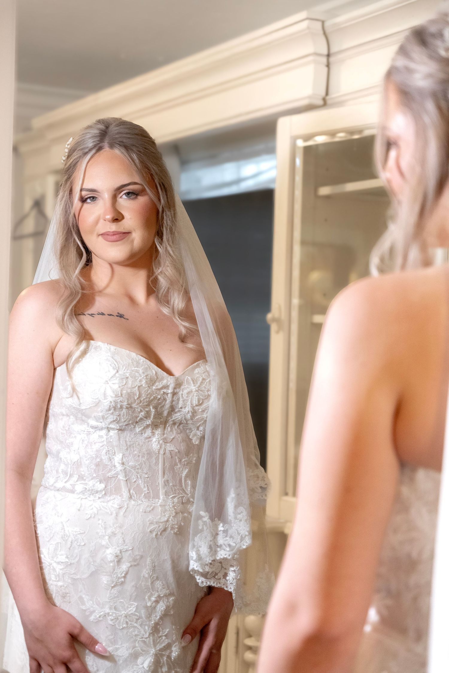 Blonde bride in lace wedding gown and veil admiring herself in a mirror.