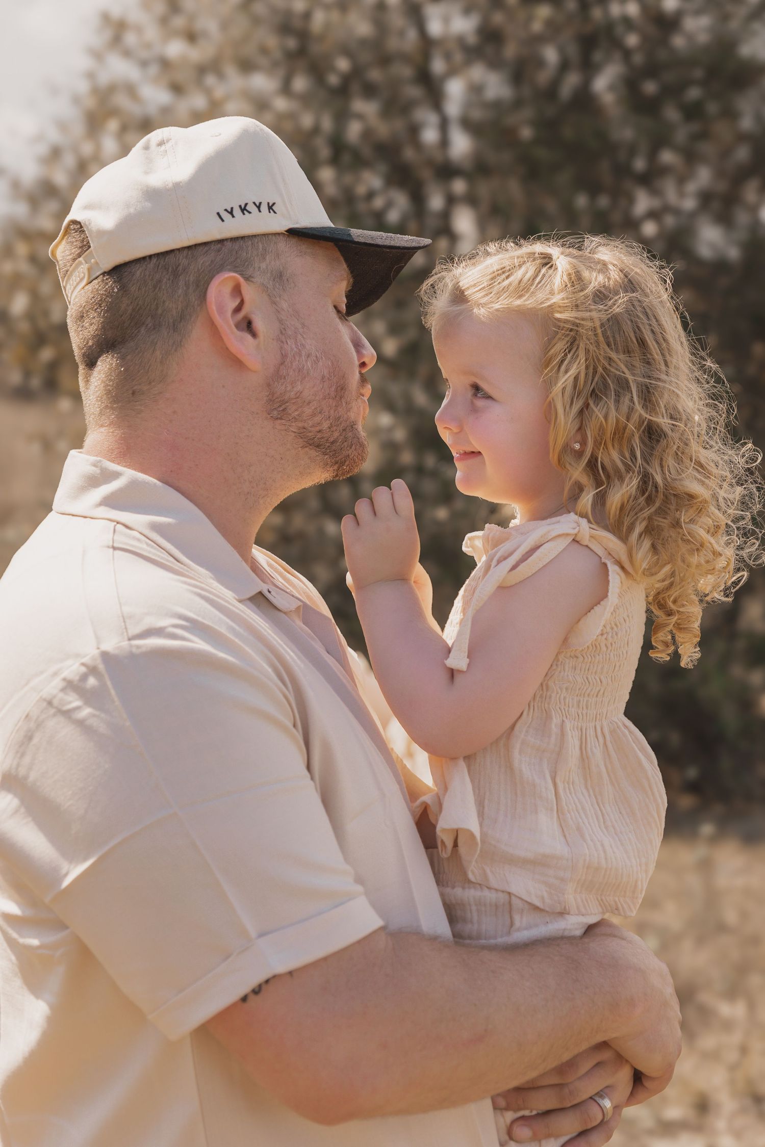 A father holds his young curly-haired daughter in a sunny outdoor setting, sharing a tender moment.
