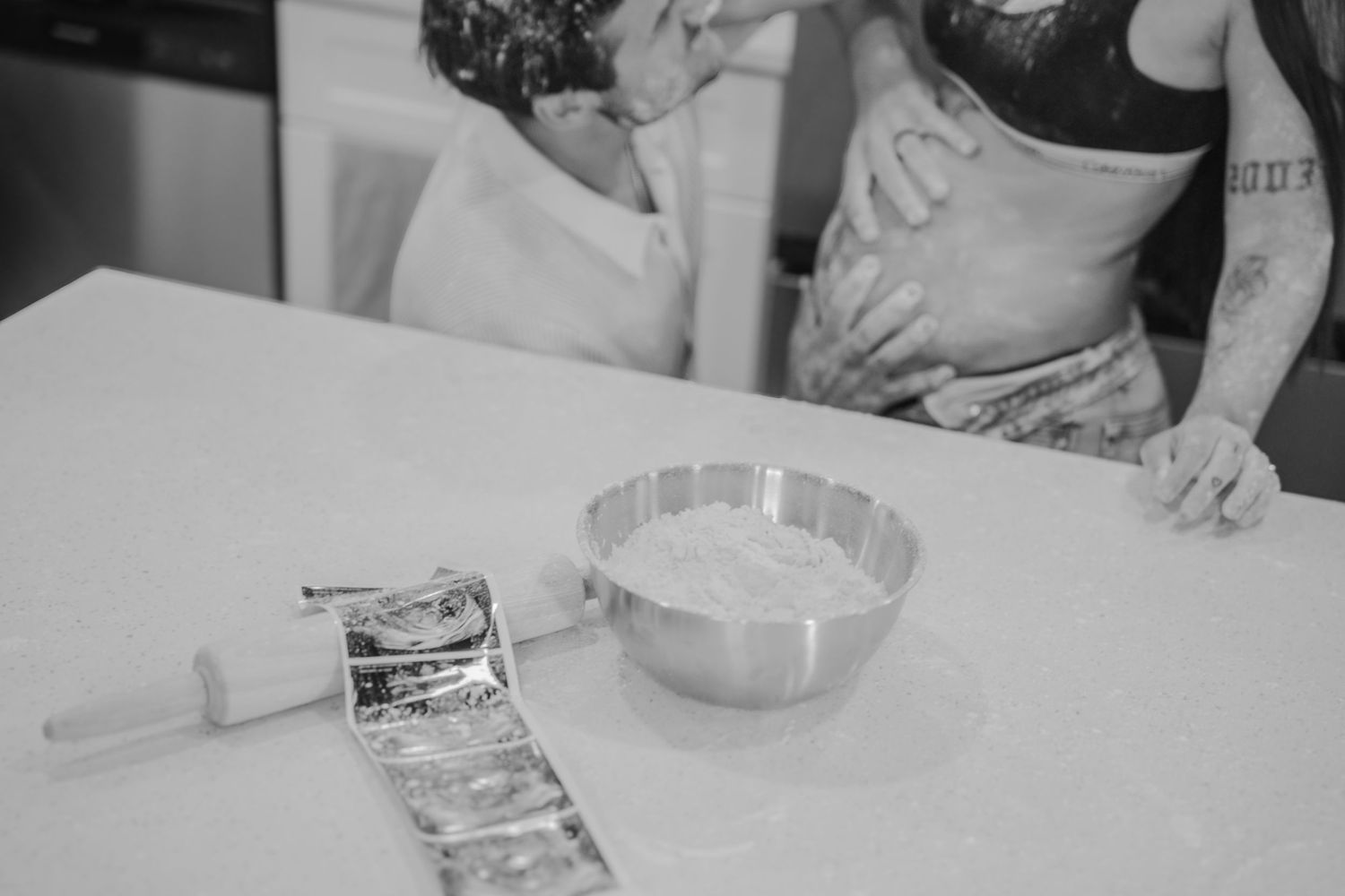Black and white photo of tattooed couple baking together at kitchen counter with mixing bowl and rolling pin.
