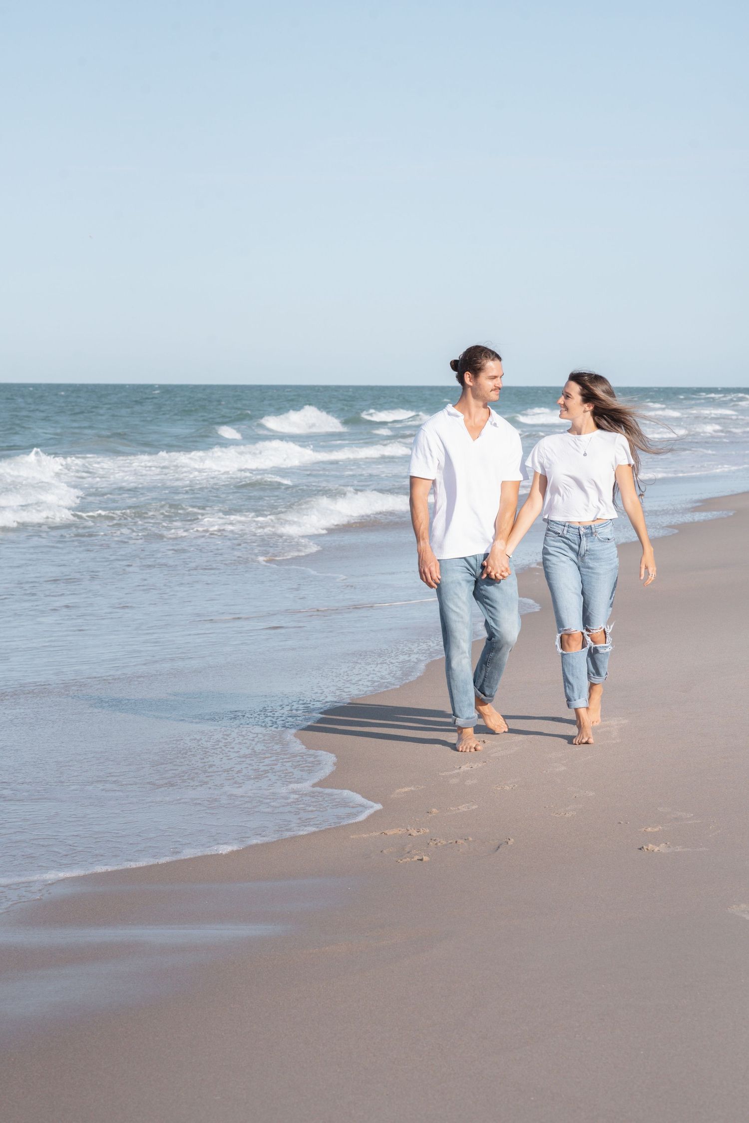 Couple in white shirts and jeans holding hands walking barefoot on a sandy beach with ocean waves.