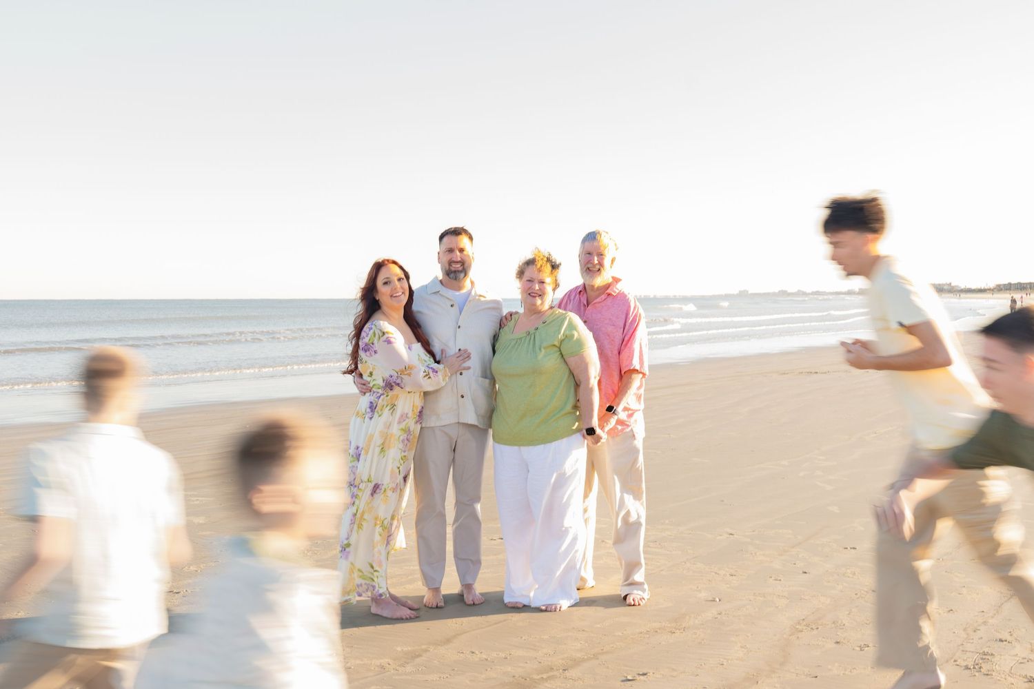 Multi-generational family posing together on a sandy beach at sunset with children playing nearby.
