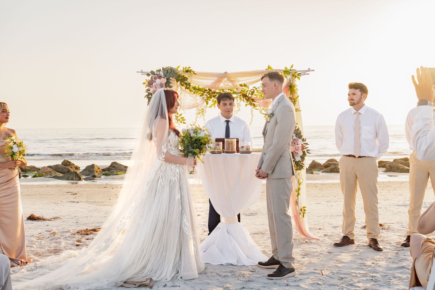Couple exchanging vows at a romantic beach wedding ceremony with floral arch and ocean backdrop at sunset.