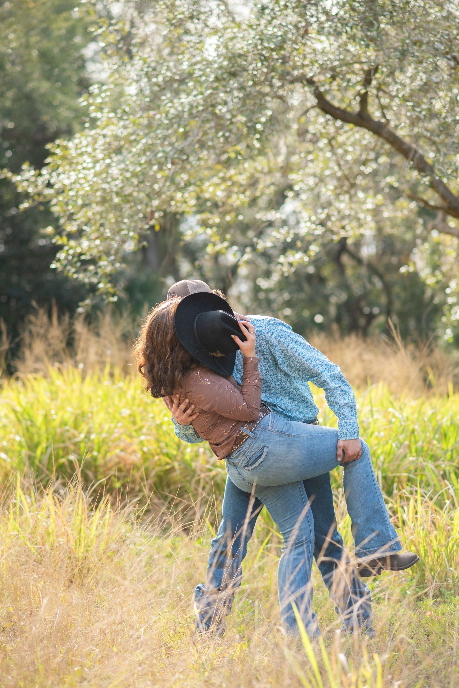 Couple embracing in a sunlit meadow, woman in blue shirt and jeans holding a black cowboy hat outdoors.