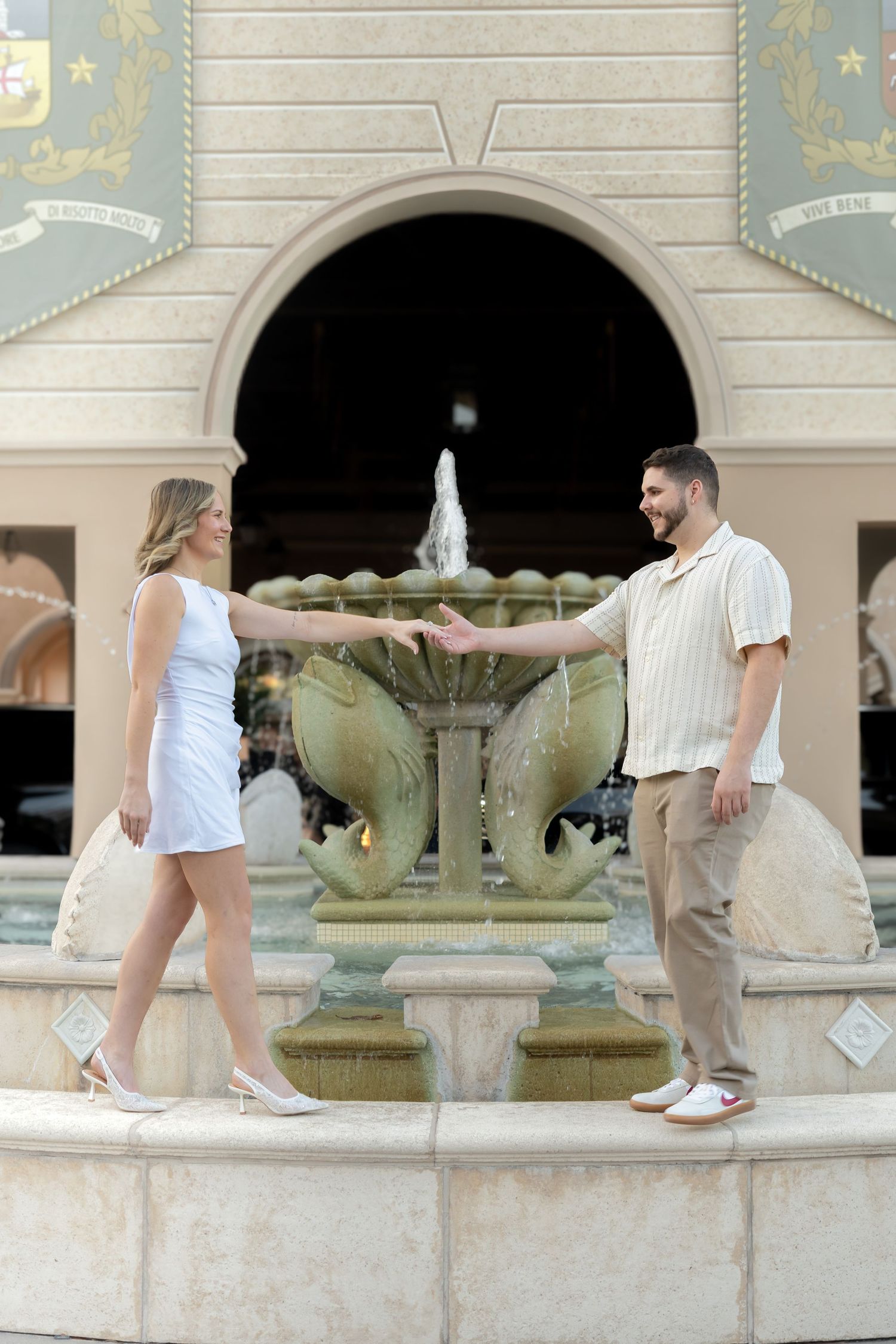 Couple holding hands by an ornate fountain with arched architecture backdrop.