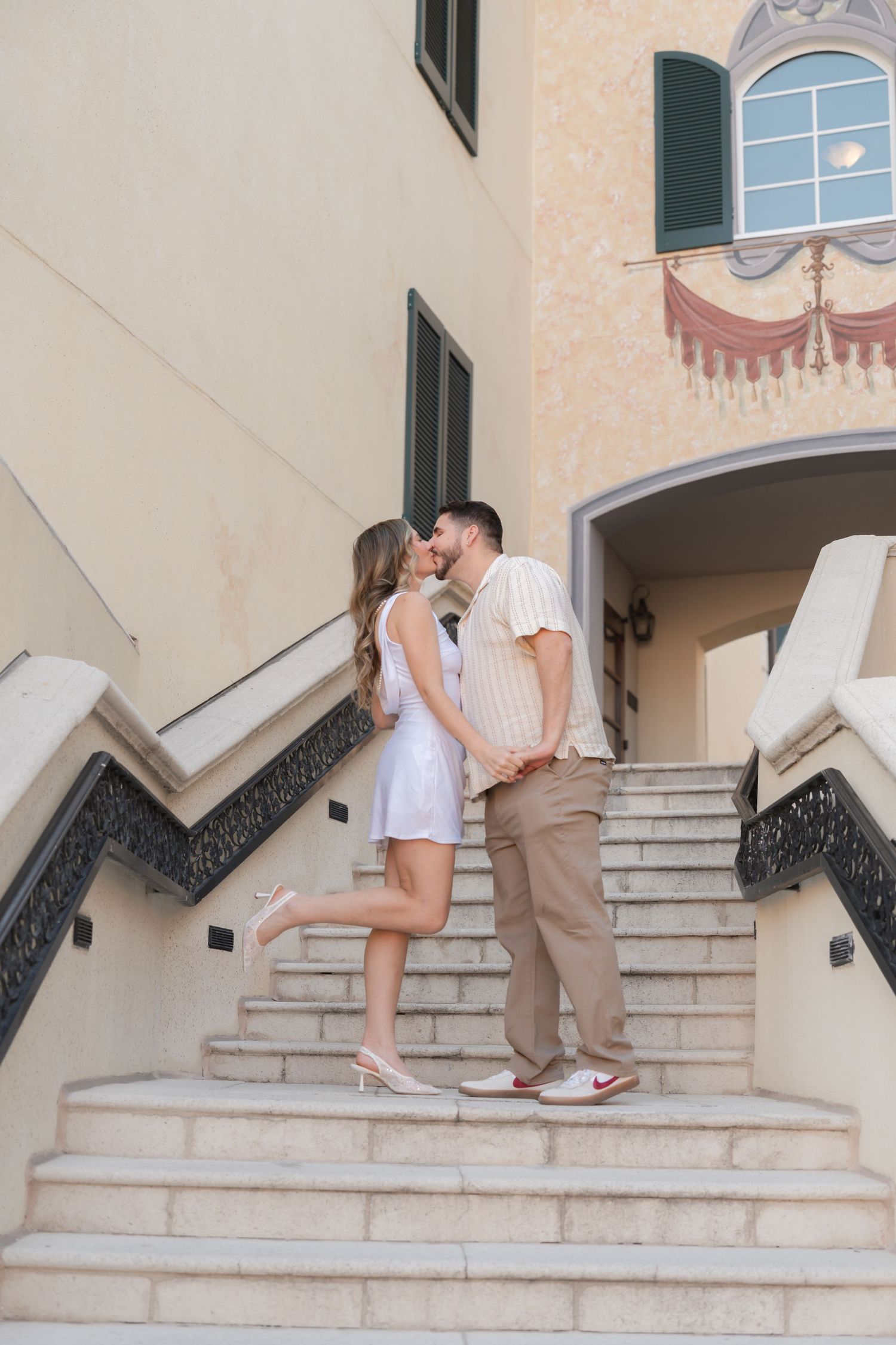 Couple sharing a romantic kiss on elegant marble stairs outside a Mediterranean-style building.