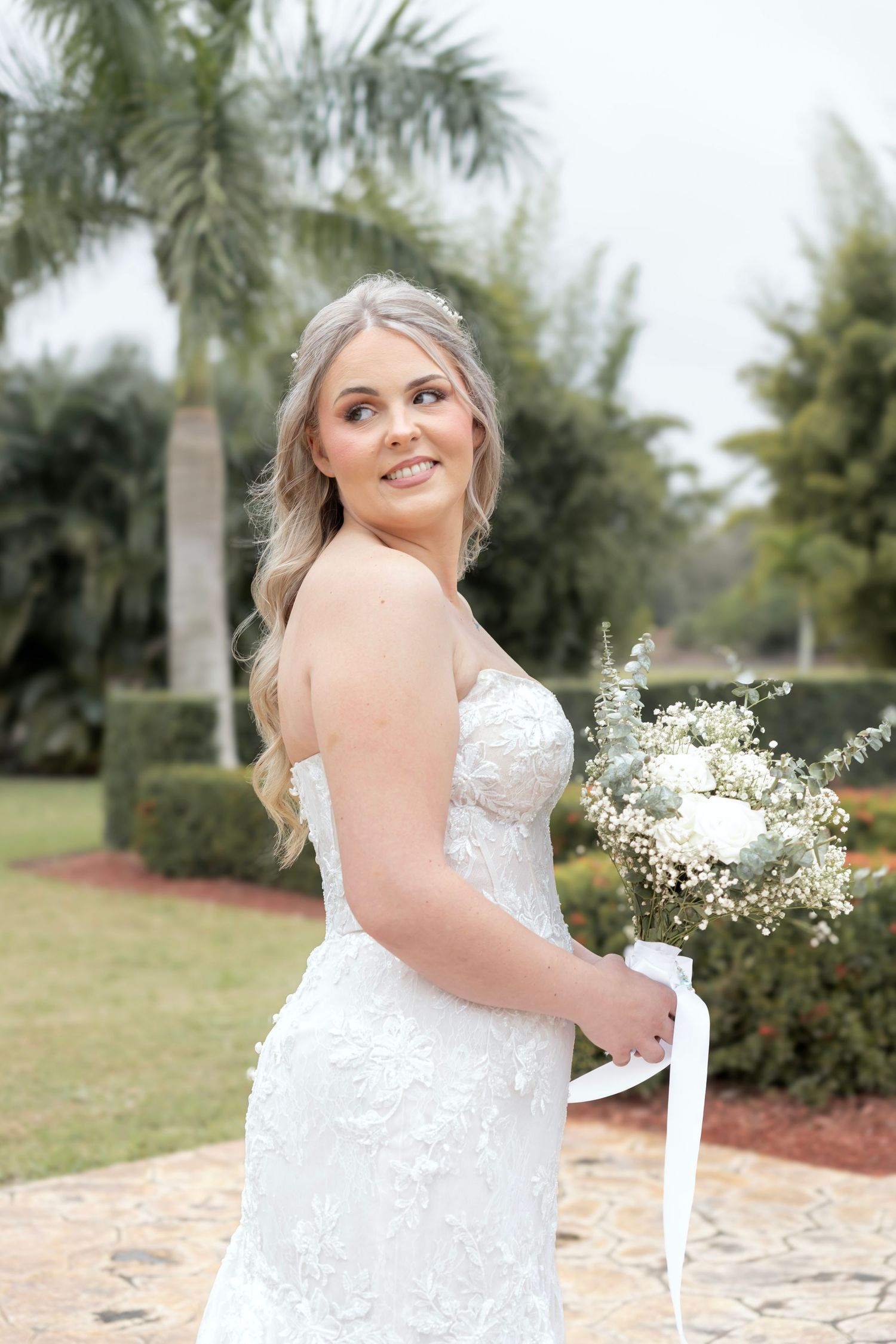Smiling bride in lace wedding gown holding white bouquet with ribbon, posing outdoors with palm trees.