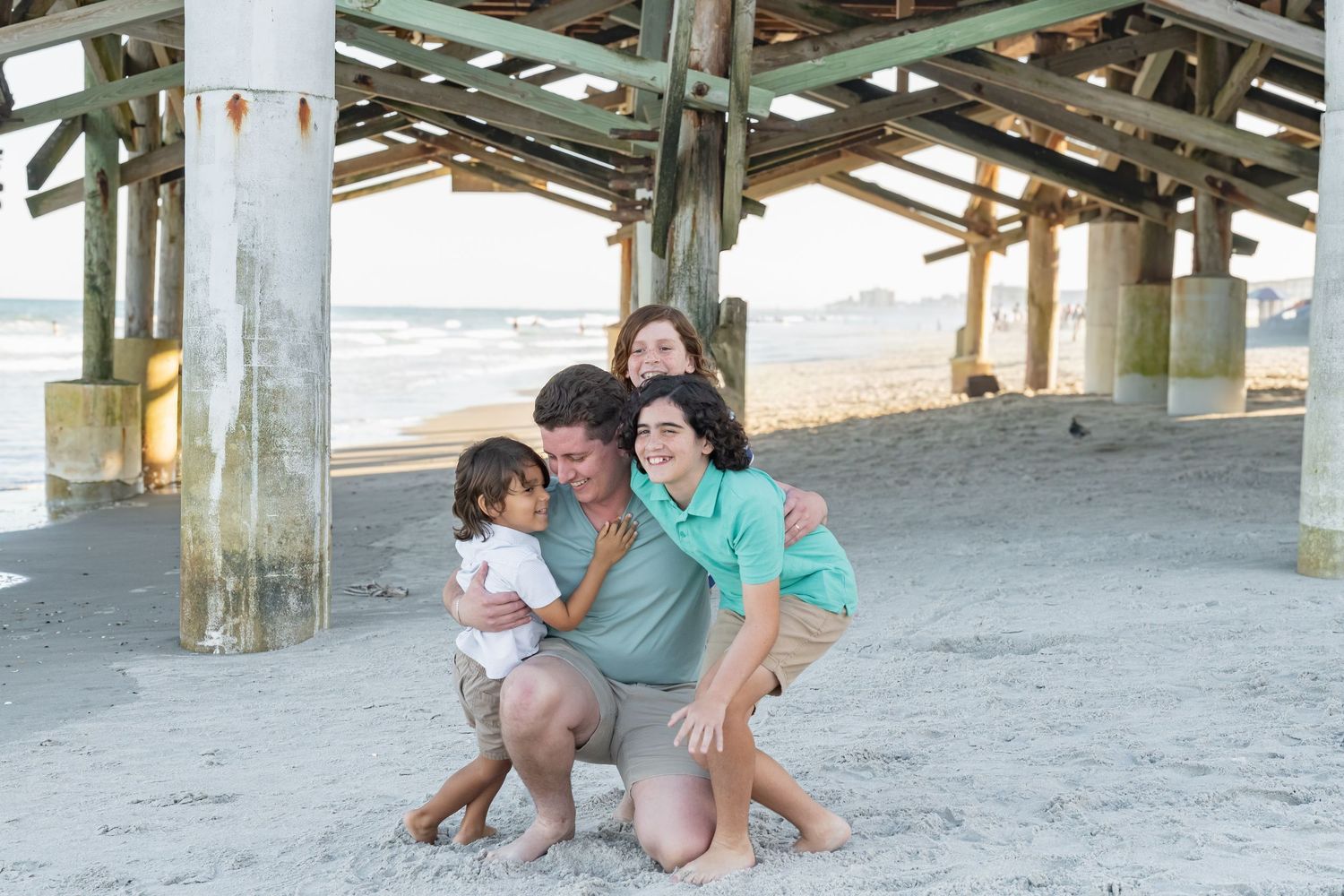 Family of four smiling and hugging on sandy beach under weathered pier structure near ocean shoreline.