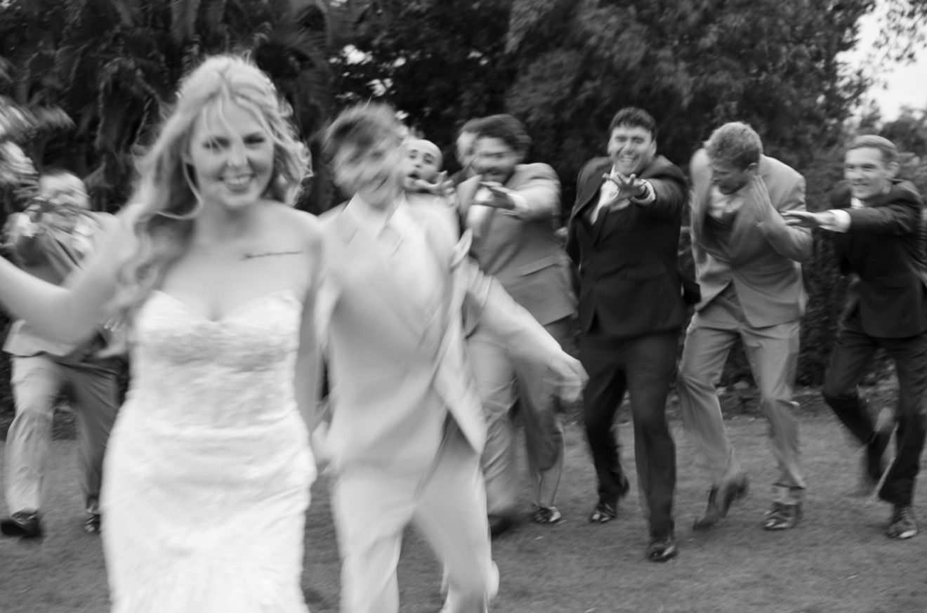 Black and white photo of joyful bride leading energetic wedding party guests across garden lawn.