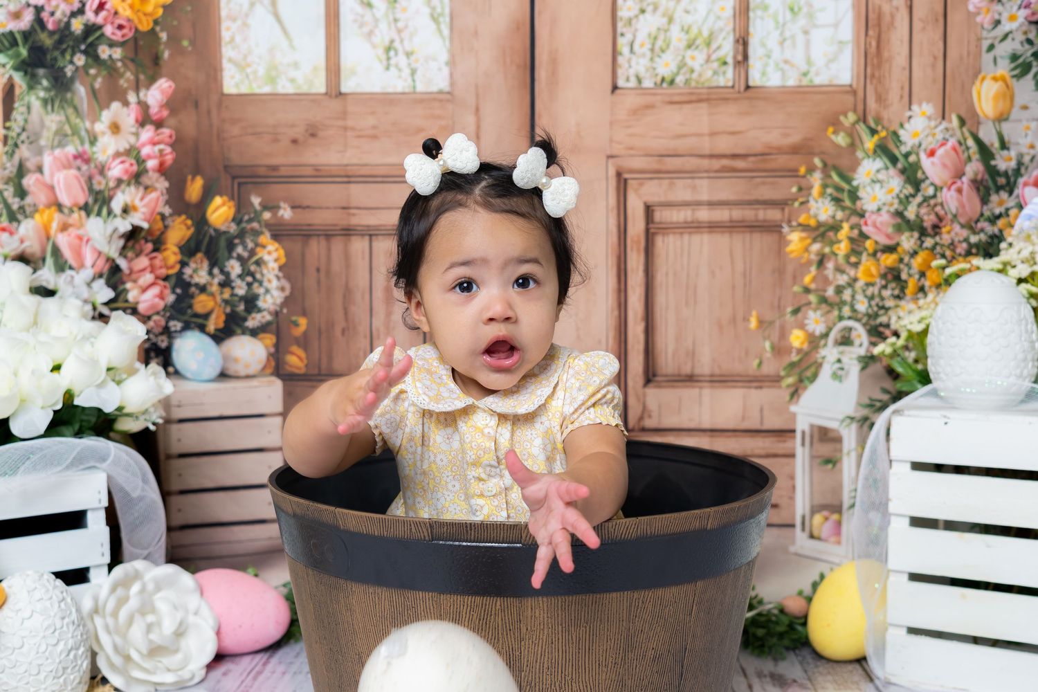 Adorable baby girl in yellow dress sits in a wooden barrel surrounded by colorful Easter floral decorations.