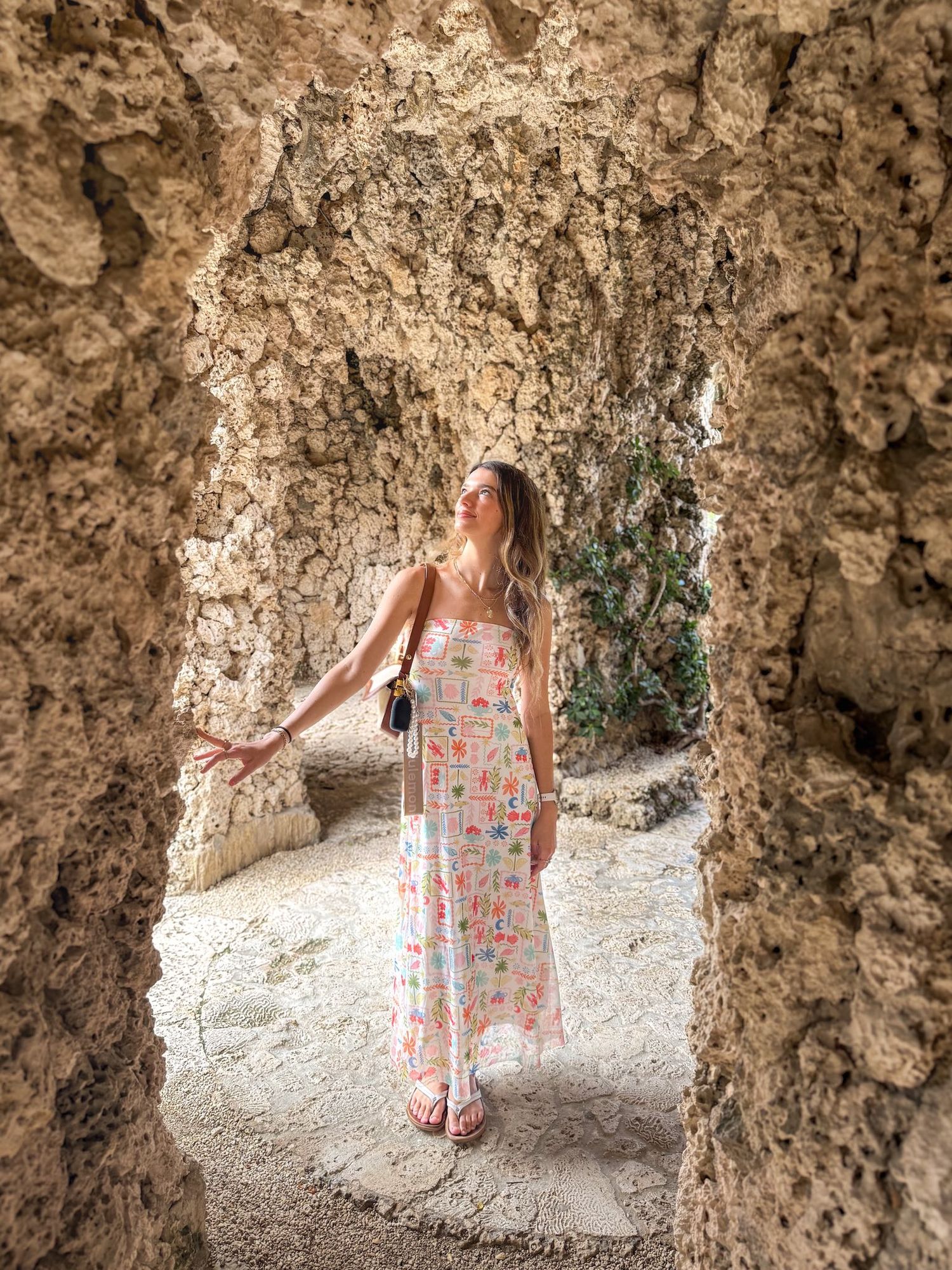 Young woman in colorful floral dress posing inside ancient stone archway with greenery background.