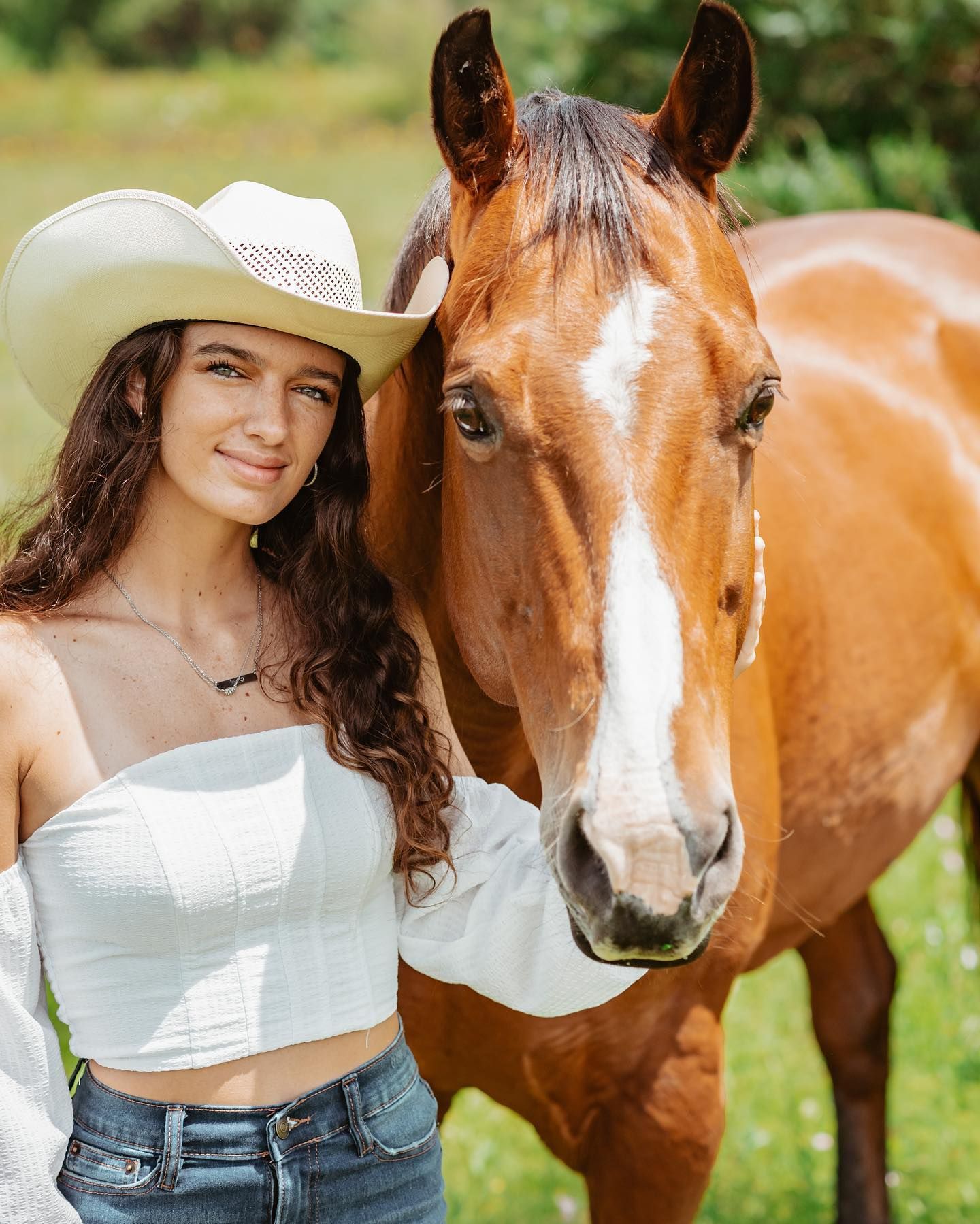 Young woman in white cowboy hat poses beside a chestnut horse in sunny green field.