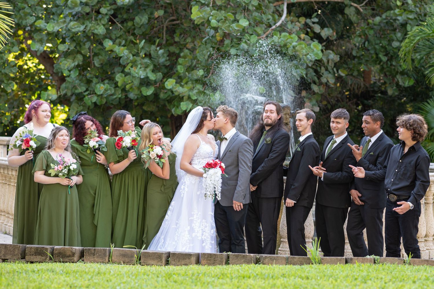 Wedding party poses in front of a fountain, bridesmaids in olive green dresses, groomsmen in black, as bride and groom share a kiss.