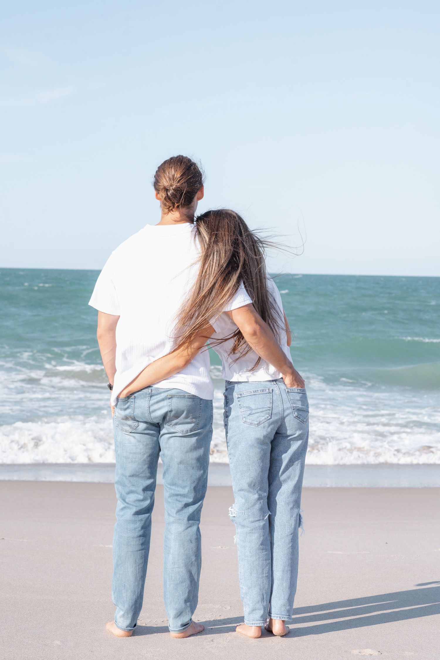 Couple in white shirts and jeans standing barefoot on a sandy beach, embracing while gazing at ocean waves.