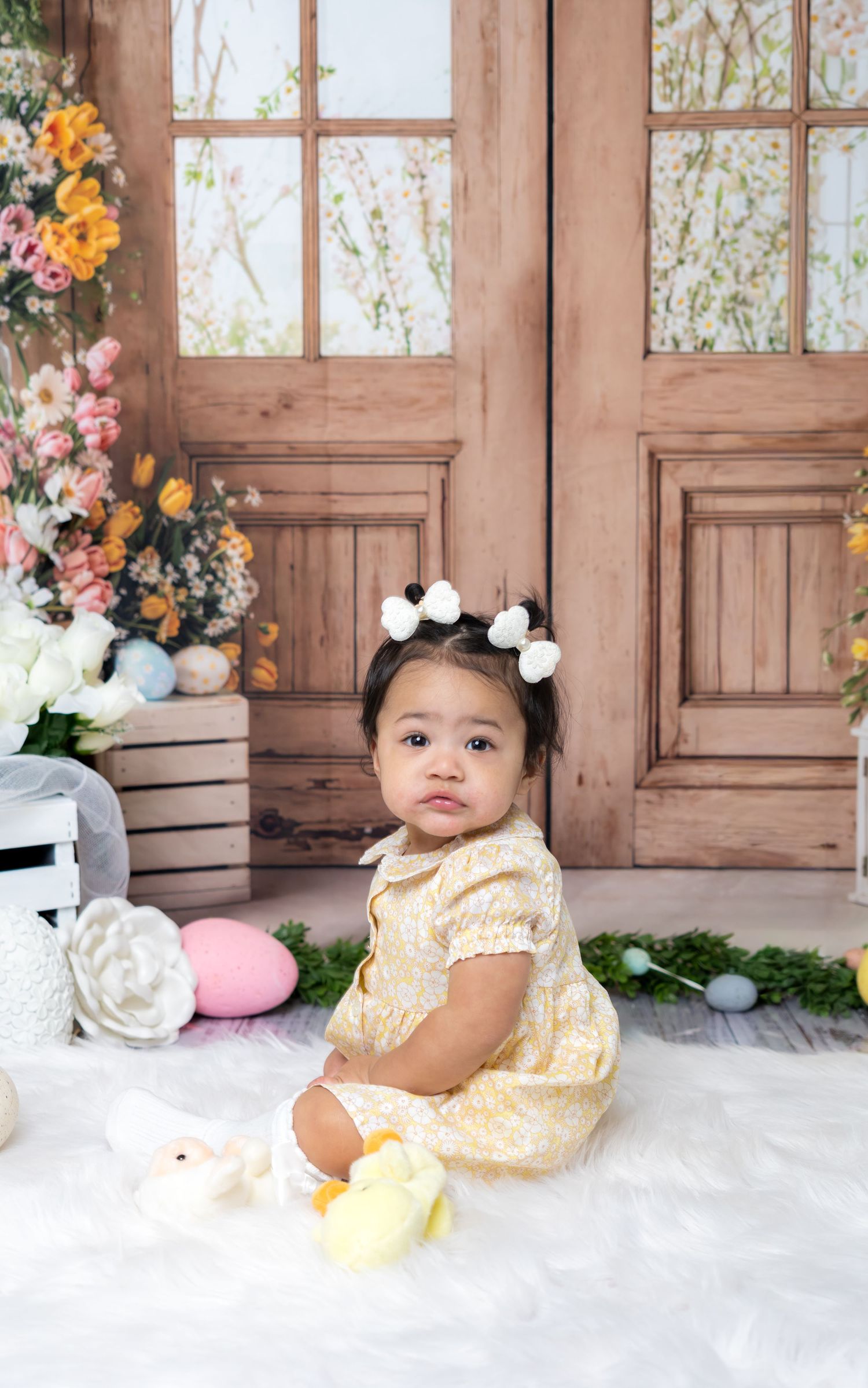 Adorable baby girl in yellow dress with white bow hair clips sits on fur rug with Easter eggs and chick toys.