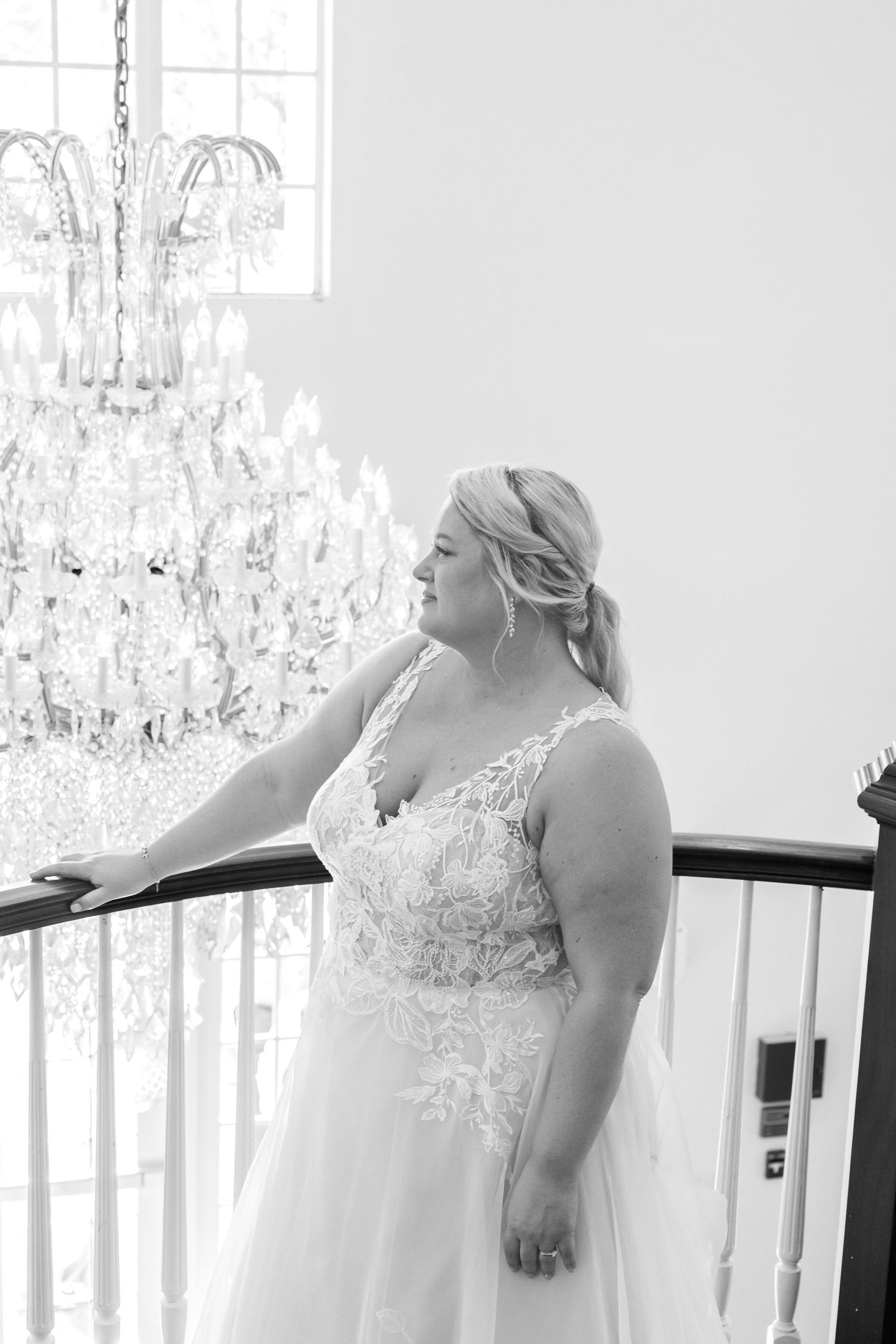 Elegant bride in lace wedding gown poses by a grand chandelier in a black and white portrait.