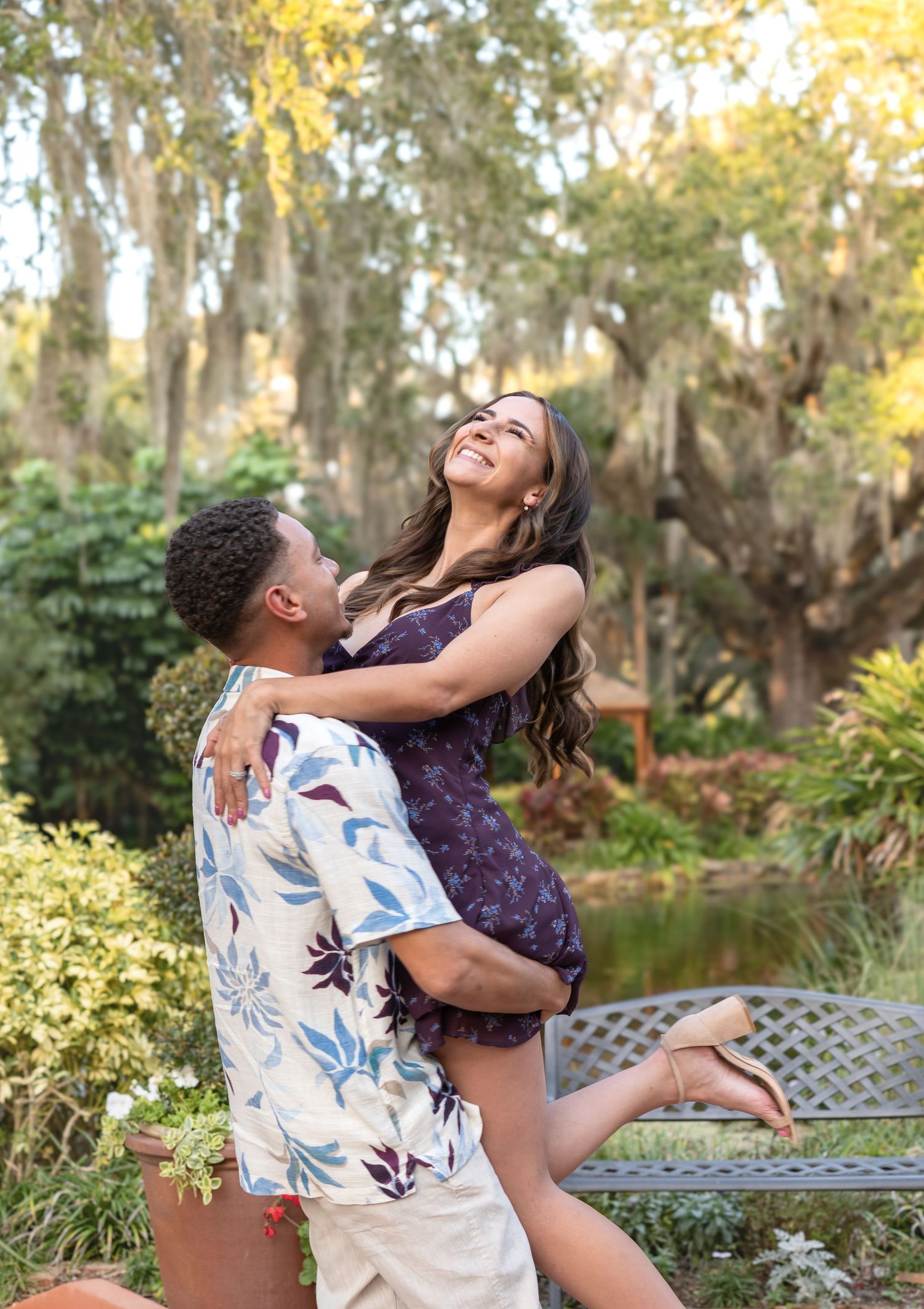 A joyful couple embraces in a lush garden, man lifting woman in floral dress, surrounded by moss-draped trees.