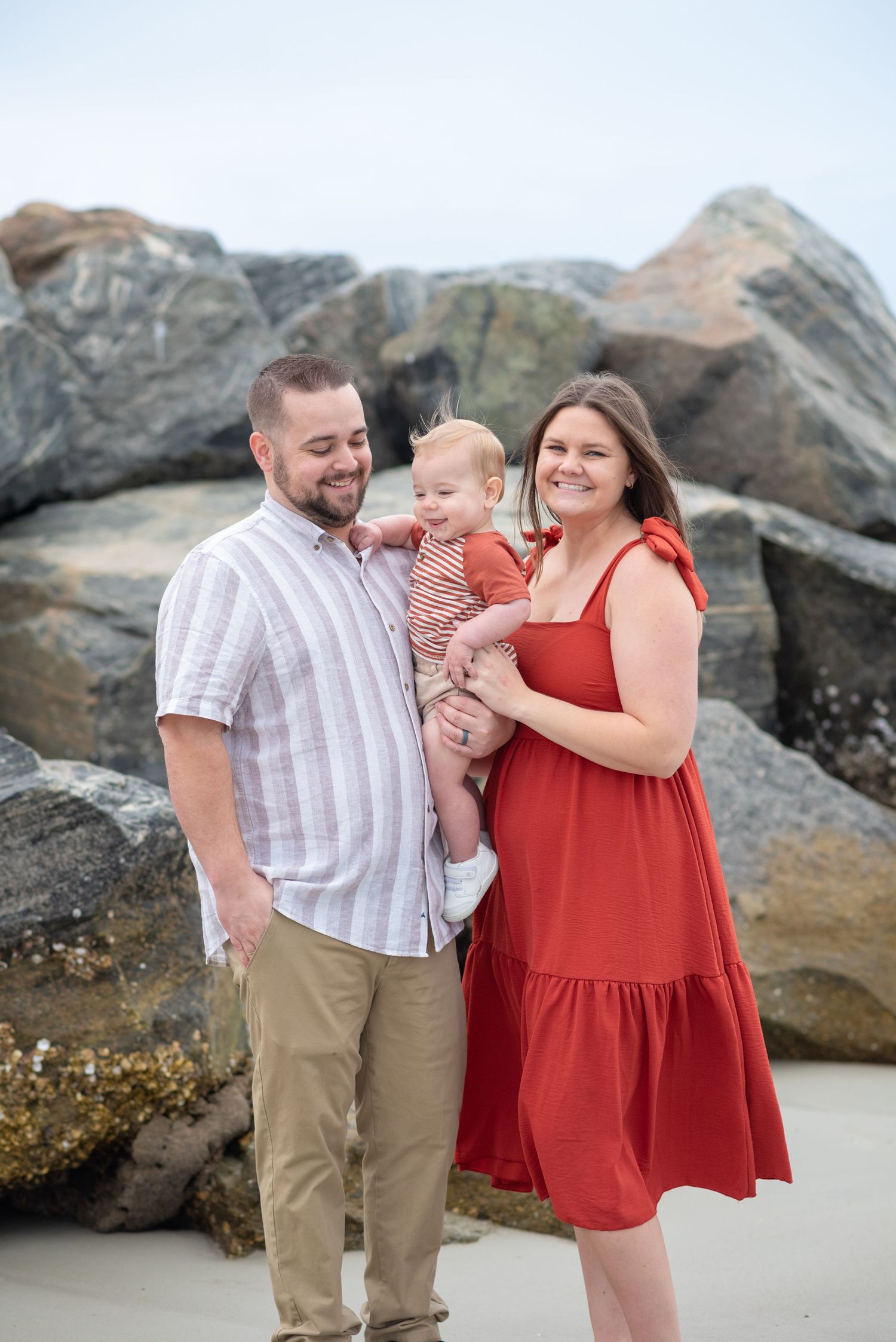 Family of three posing on rocky beach, parents smiling at baby in red outfits.