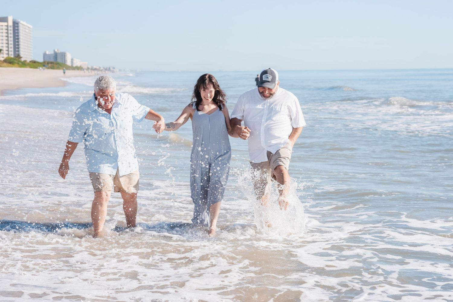 Three people laughing and splashing through ocean waves together on a sunny beach day.