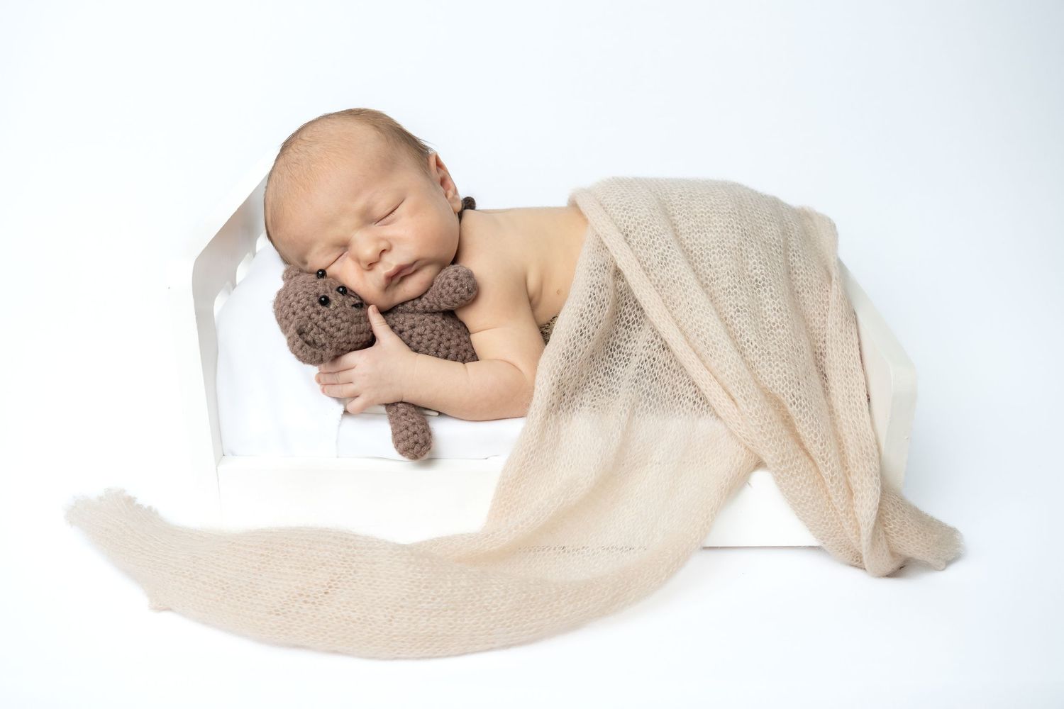 Sleeping newborn baby wrapped in beige blanket hugging brown knitted teddy bear on white background.