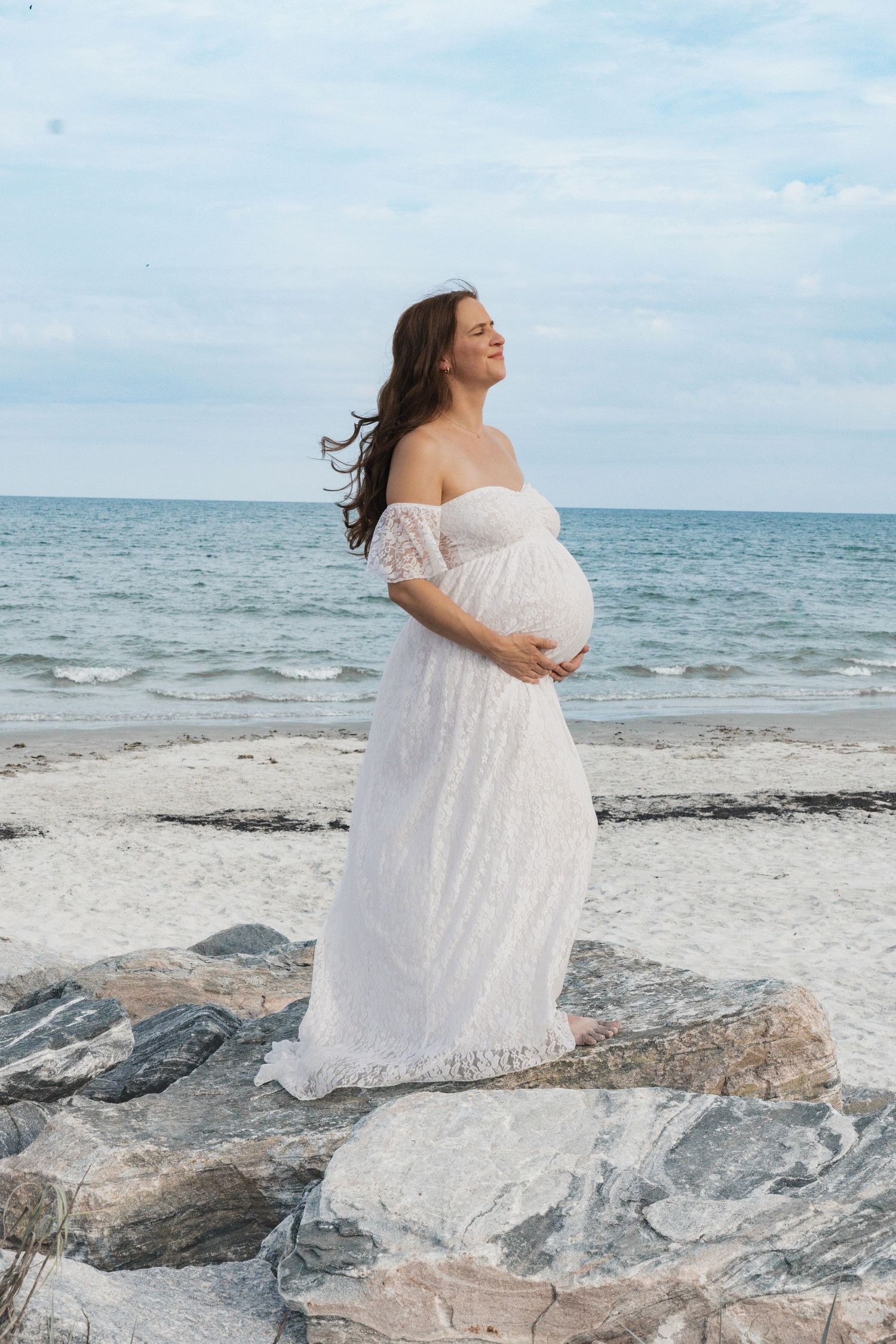 Pregnant woman in white lace off-shoulder gown stands on rocky beach, cradling belly with ocean backdrop.
