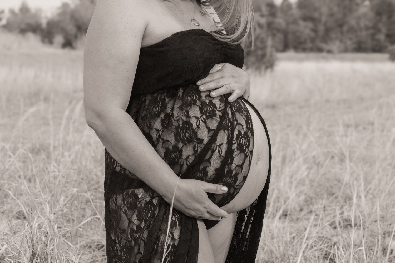 Black and white maternity photo of pregnant woman in lace dress cradling baby bump outdoors in field.