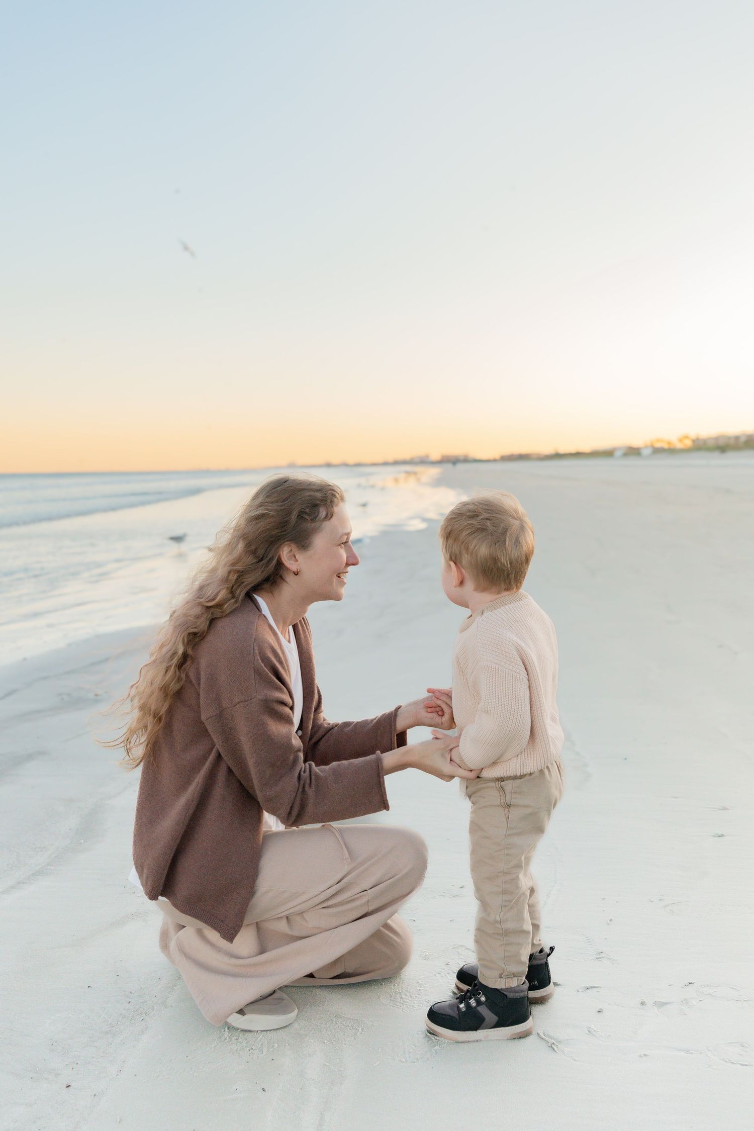 A mother crouches to talk with her toddler son on a white sand beach at sunset, both dressed in neutral tones.