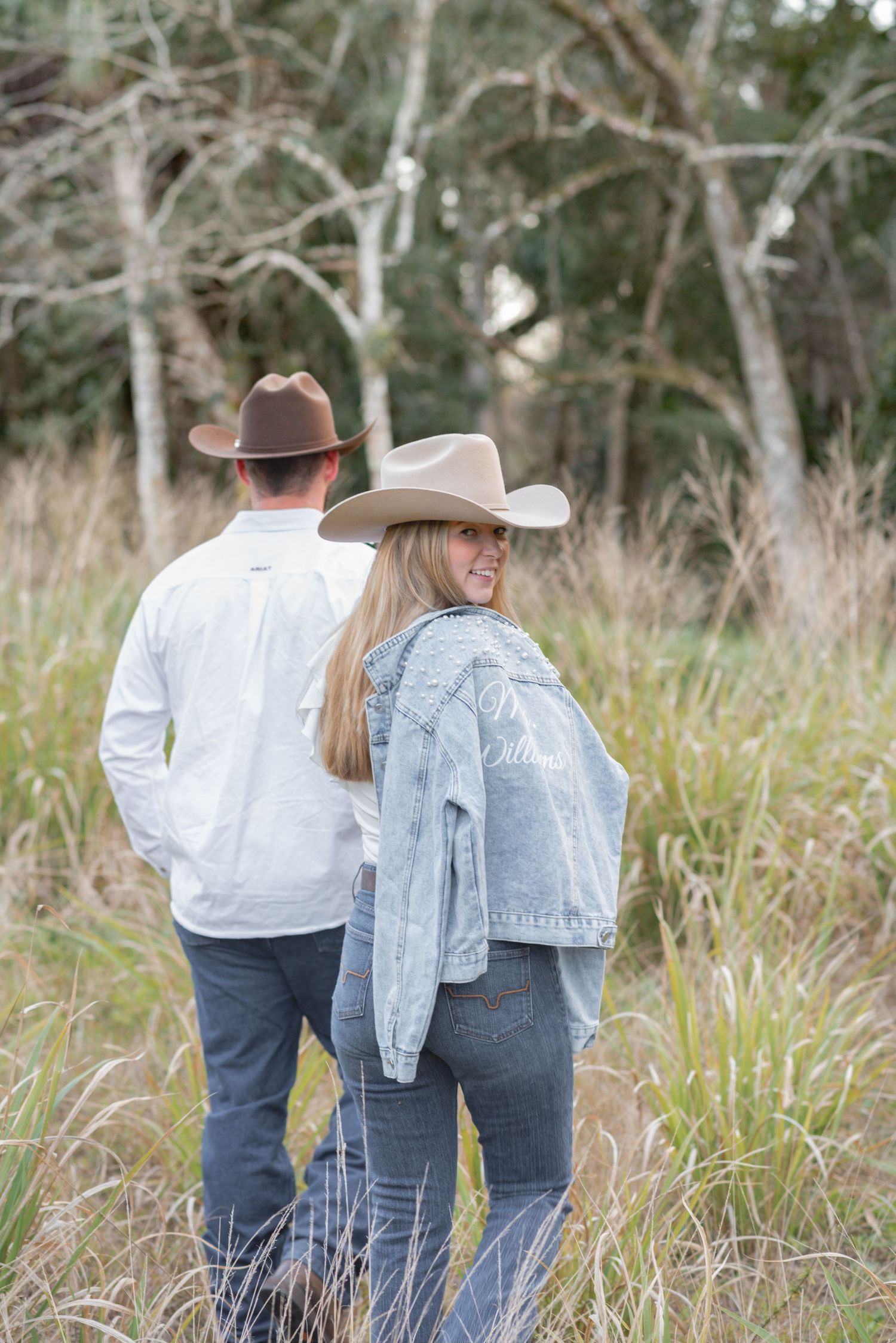 A couple in cowboy hats walk through tall grass, the woman wearing a custom Williams denim jacket, smiling back.