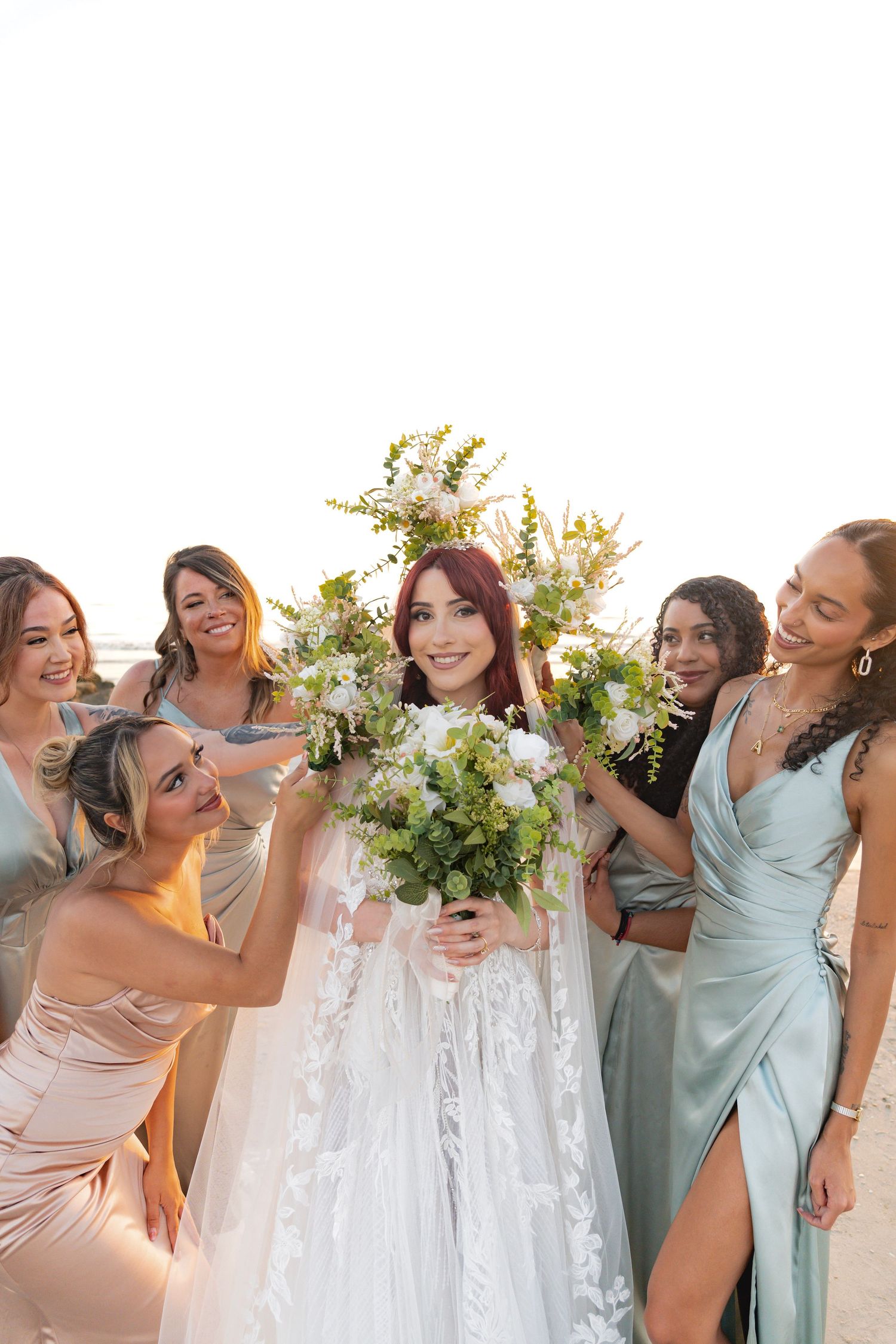 Bride in white lace gown holds large floral bouquet surrounded by smiling bridesmaids in sage and champagne dresses.