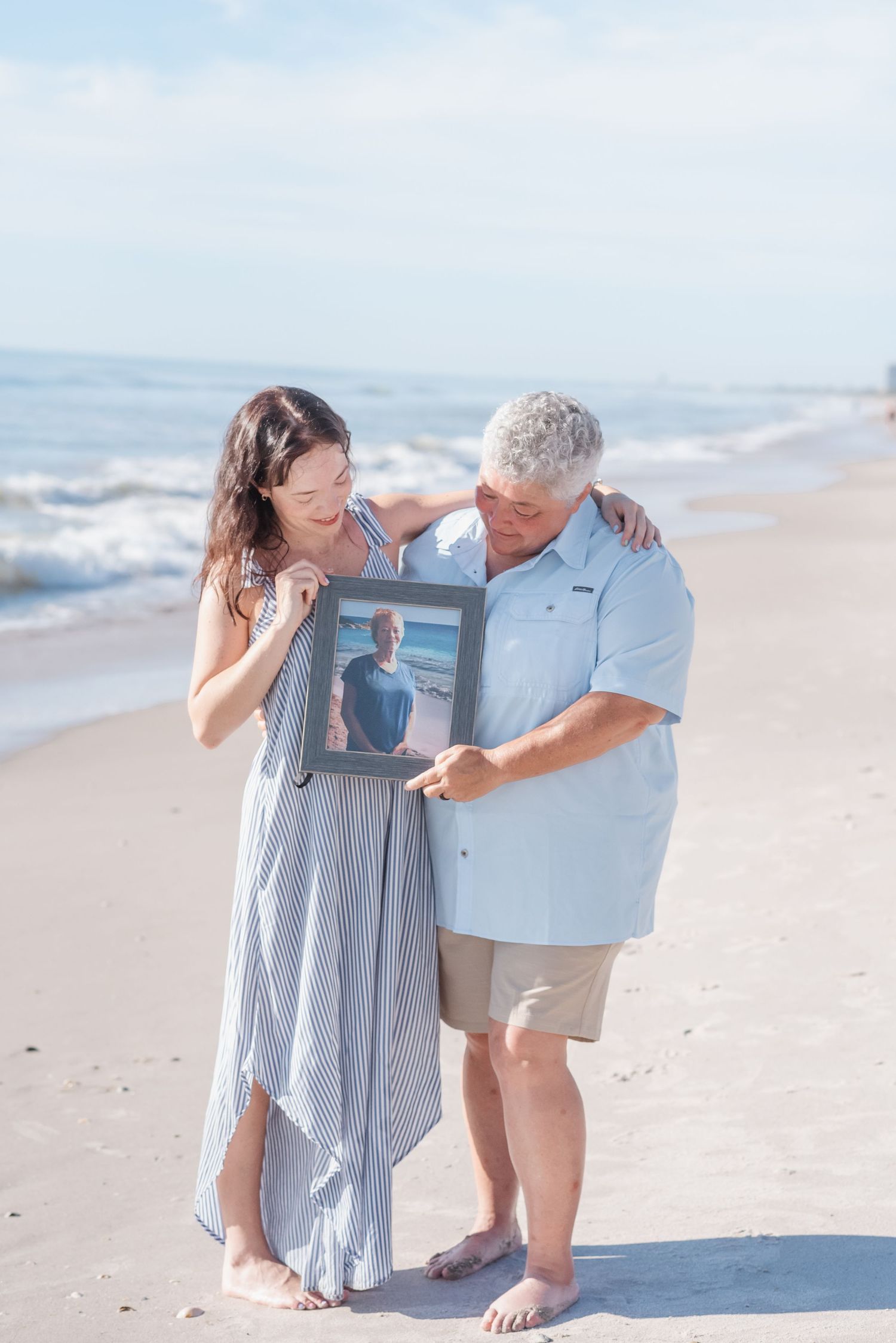 Couple standing barefoot on beach holding framed photo of loved one, ocean waves in background.