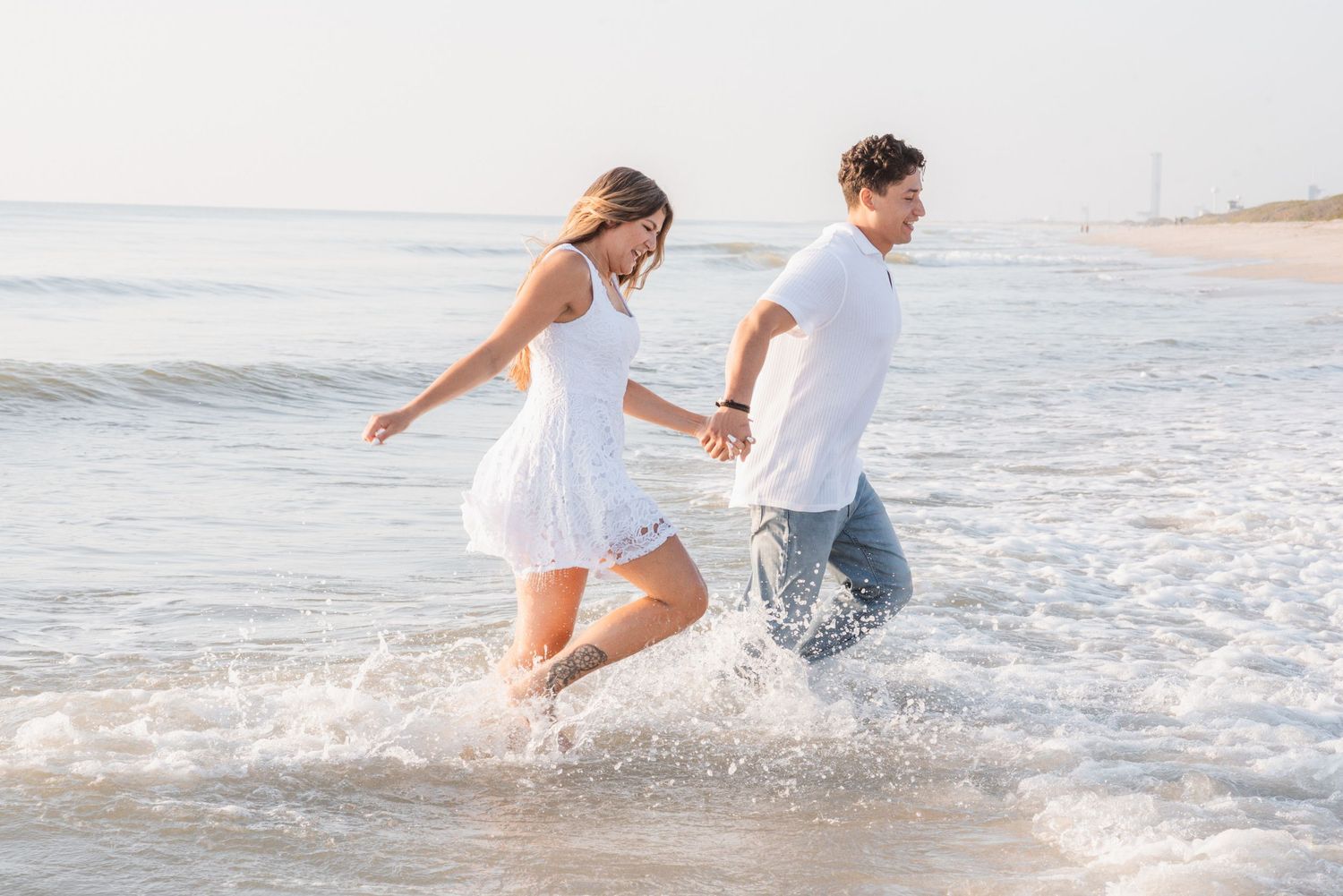 Couple holding hands running through ocean waves on a sunny beach, woman in white dress and man in white shirt.