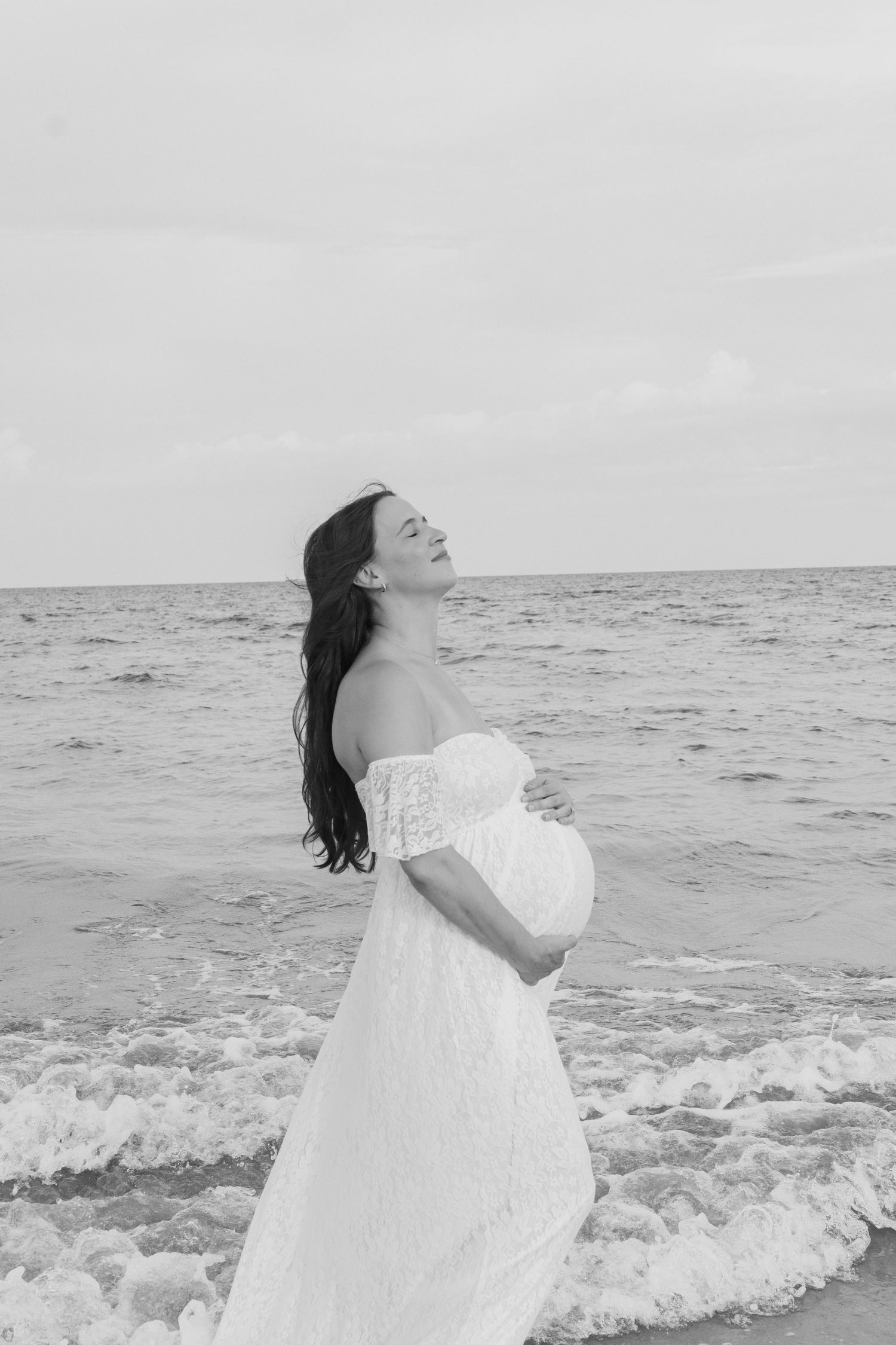 Black and white photo of pregnant woman in white lace dress cradling belly on beach with ocean waves behind her.