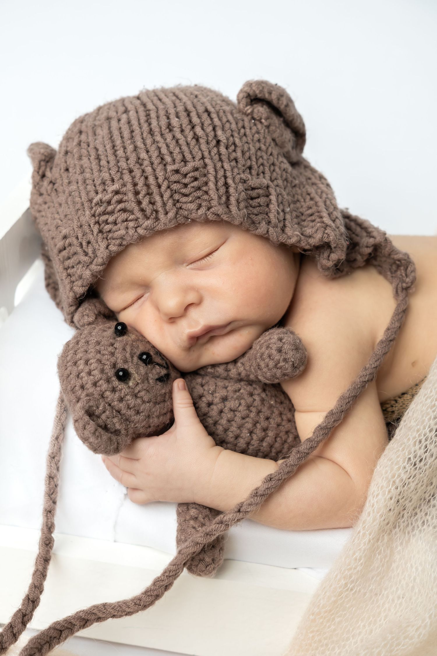 Sleeping newborn baby wearing a knit brown bear hat holding a matching crochet teddy bear toy in a white crib.