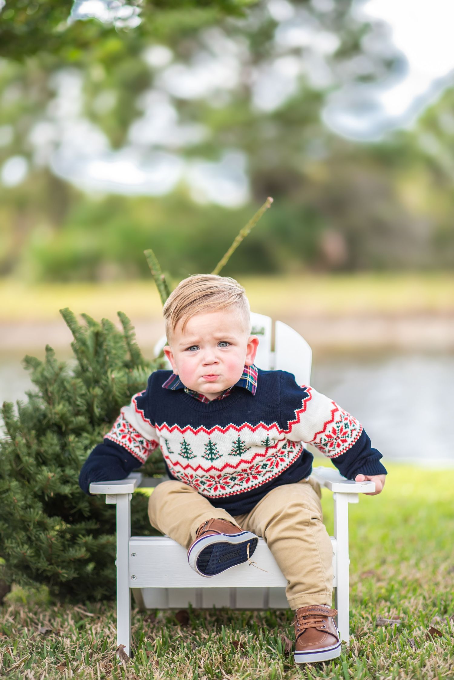 Toddler boy in festive Fair Isle sweater sitting in white Adirondack chair beside small Christmas tree outdoors.