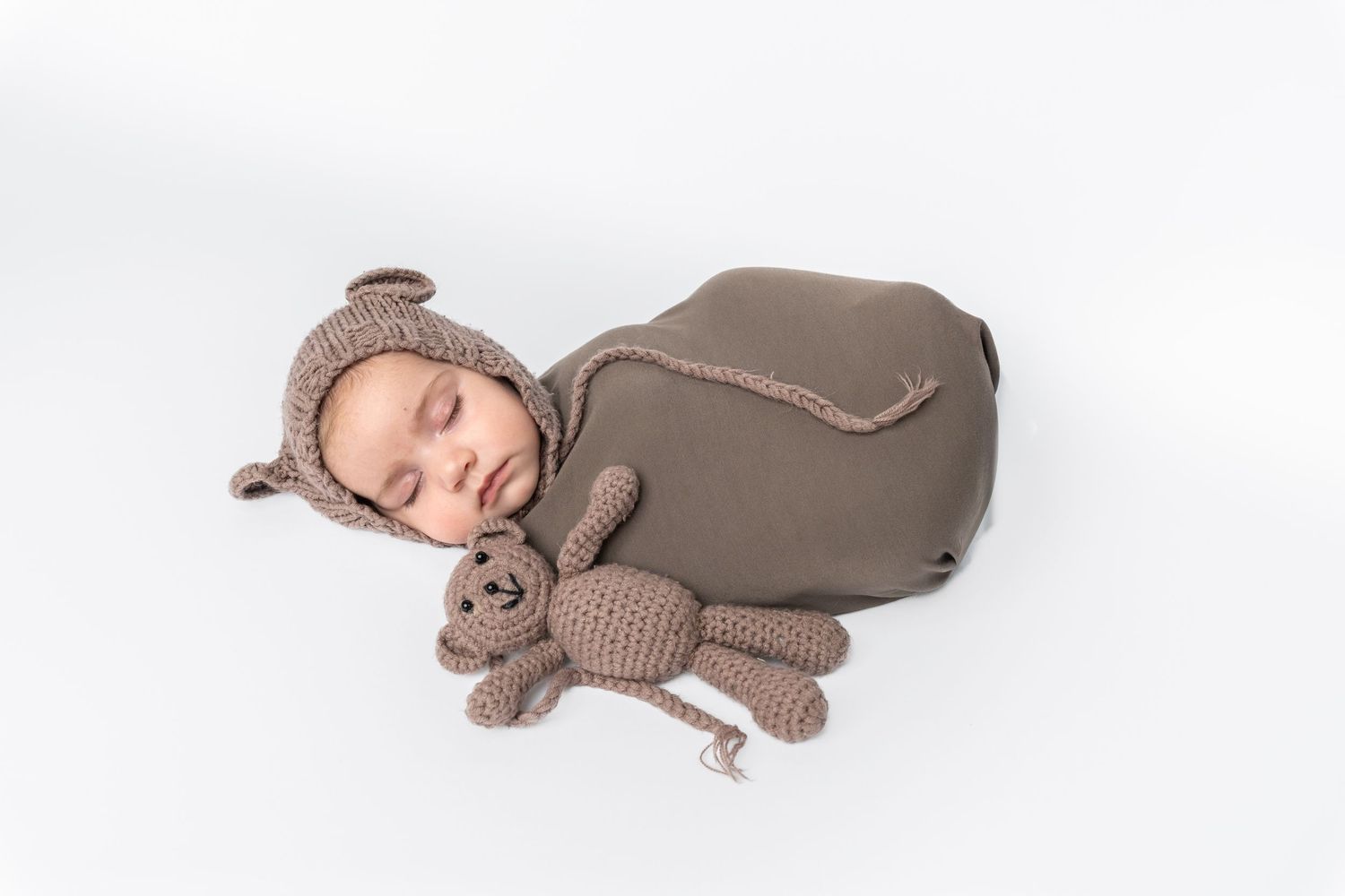 Newborn baby sleeping in brown bear swaddle wrap with matching crochet teddy bear on white background.