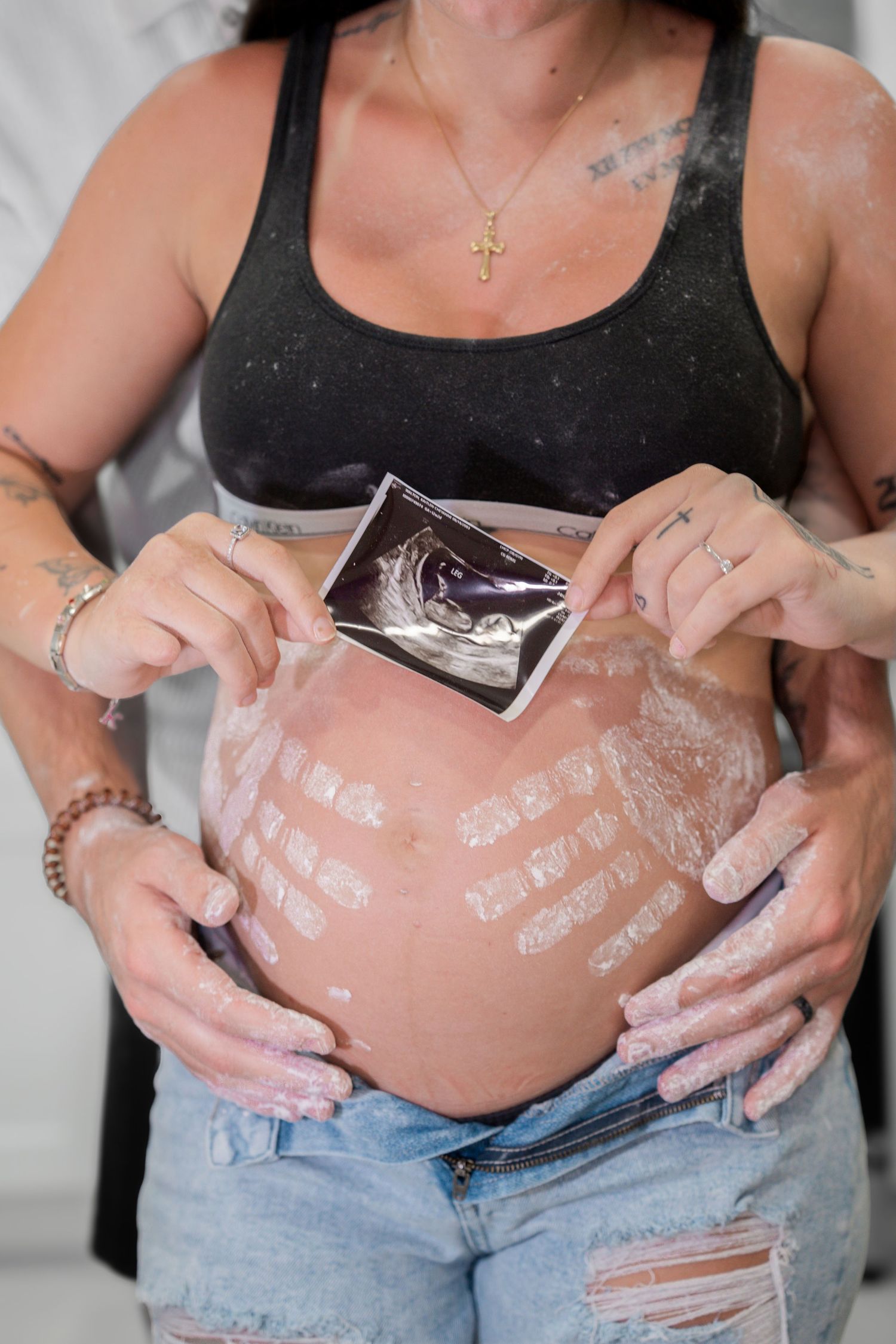 Pregnant woman holding ultrasound photo over baby bump with painted stretch marks, wearing jeans and black crop top.