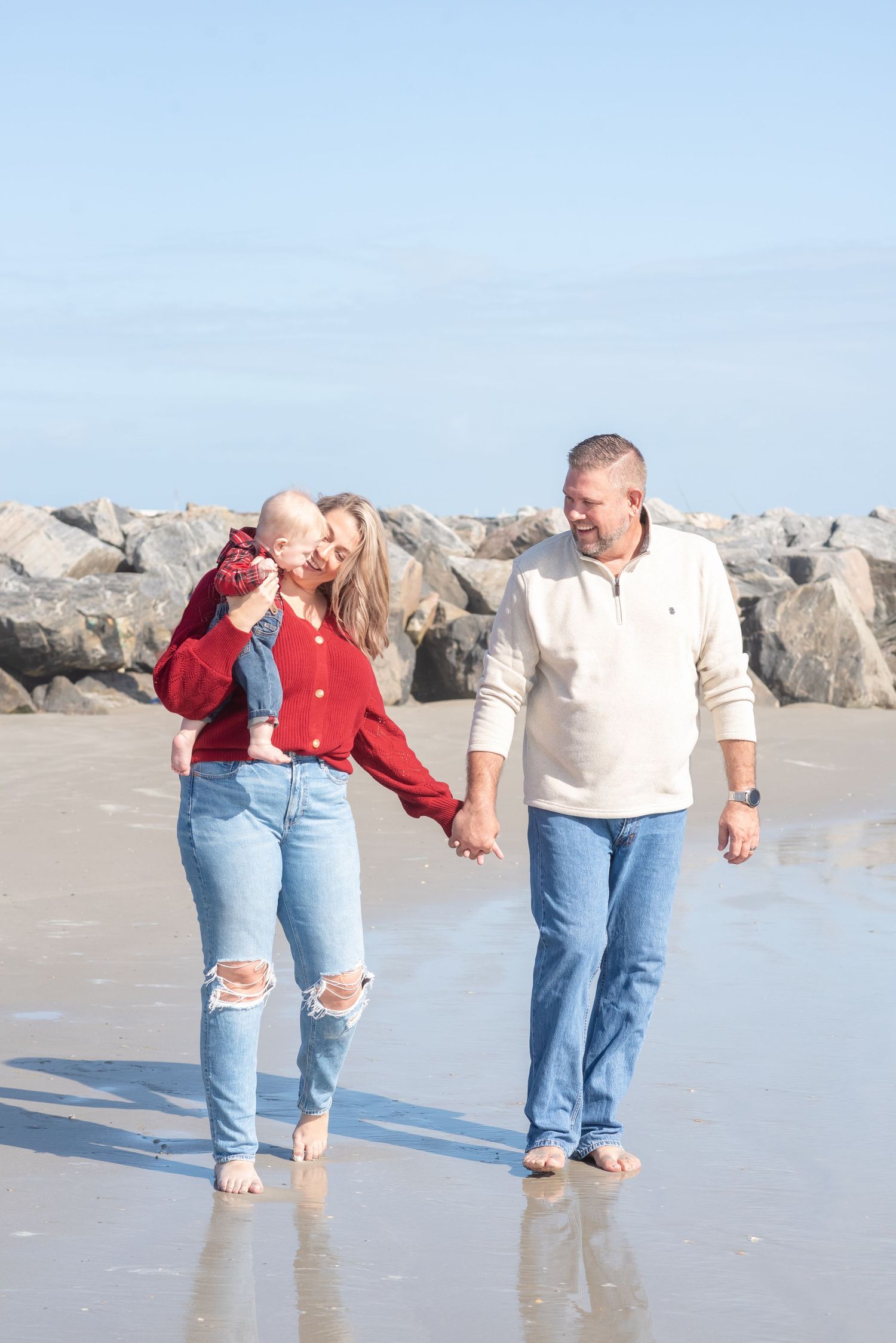 Family of three walking barefoot on a beach, mother holding baby, father smiling, rocky jetty in background.